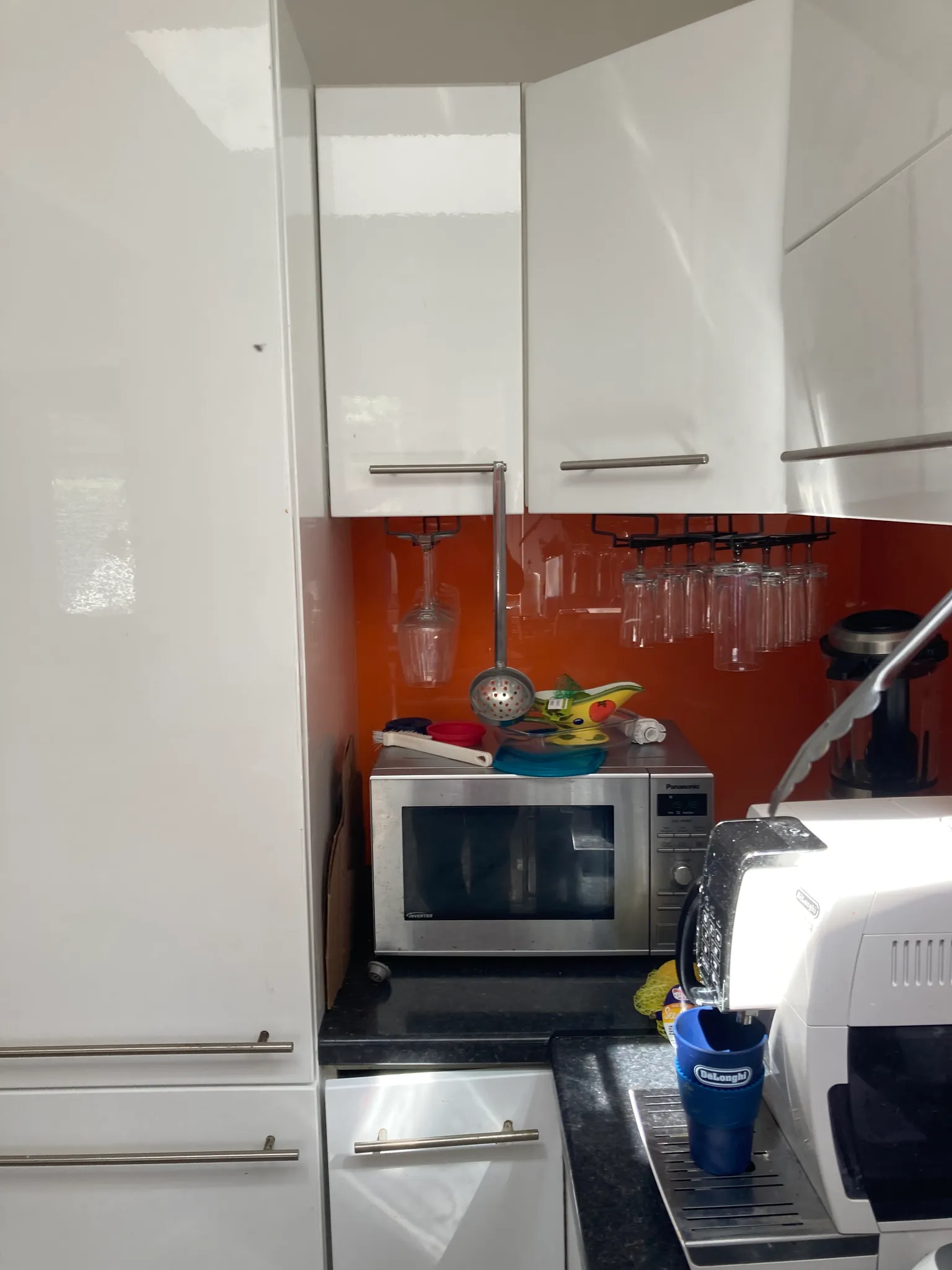 Kitchen area with white upper cabinets, an orange backsplash, a microwave on the counter, hanging wine glasses and a coffee machine.