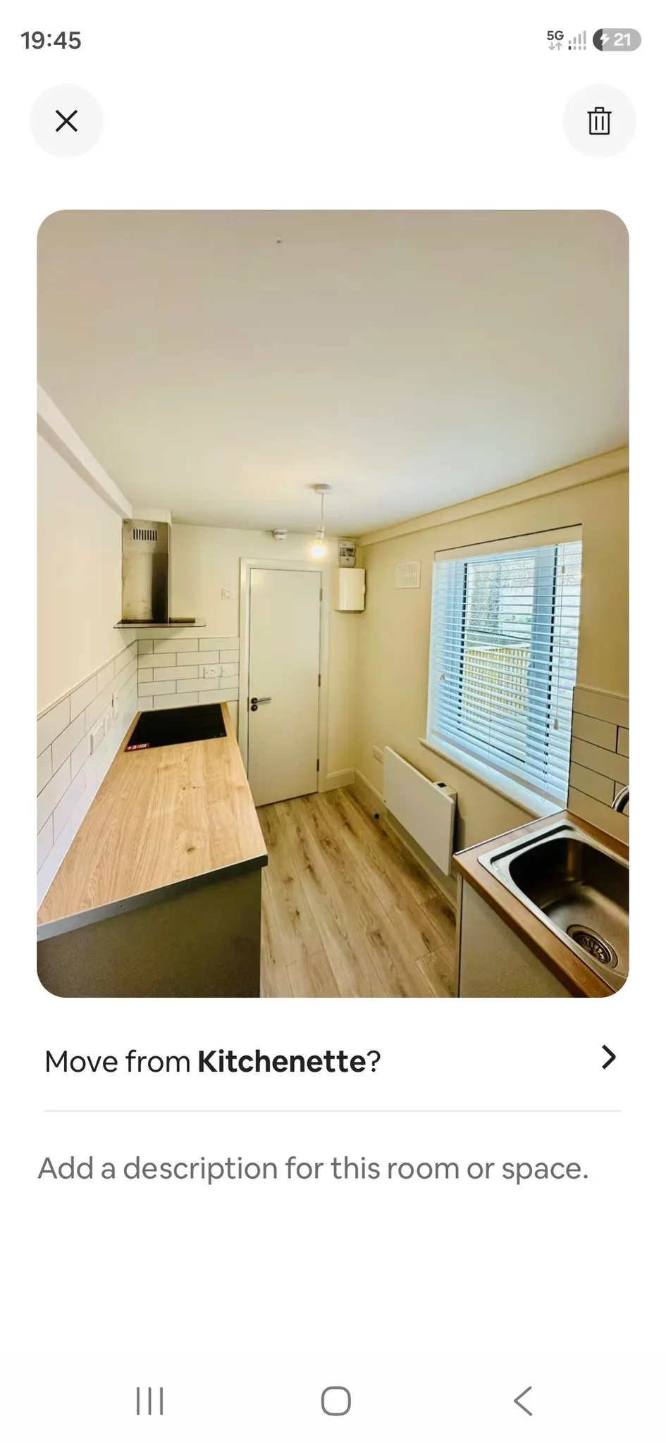 Narrow kitchen with wooden countertop, induction hob, stainless sink, white tiled backsplash, window with blinds, radiator, and a closed door at the end.