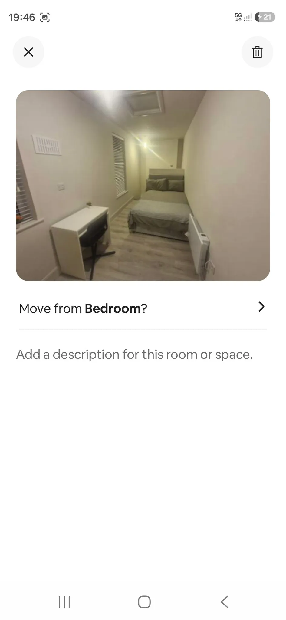 Narrow bedroom with a bed against the far wall, small desk and chair near a window with blinds, radiator on the right, and overhead light.