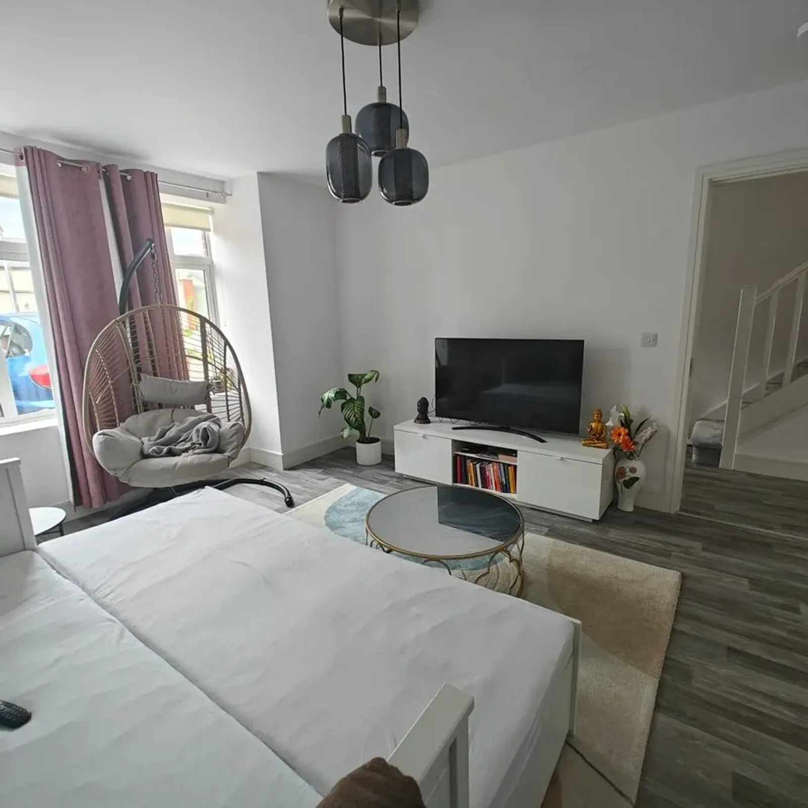 Bedroom with a double bed, wall TV on a low media console, round glass coffee table, hanging rattan chair by a window with curtains, potted plant, and pendant ceiling lights.