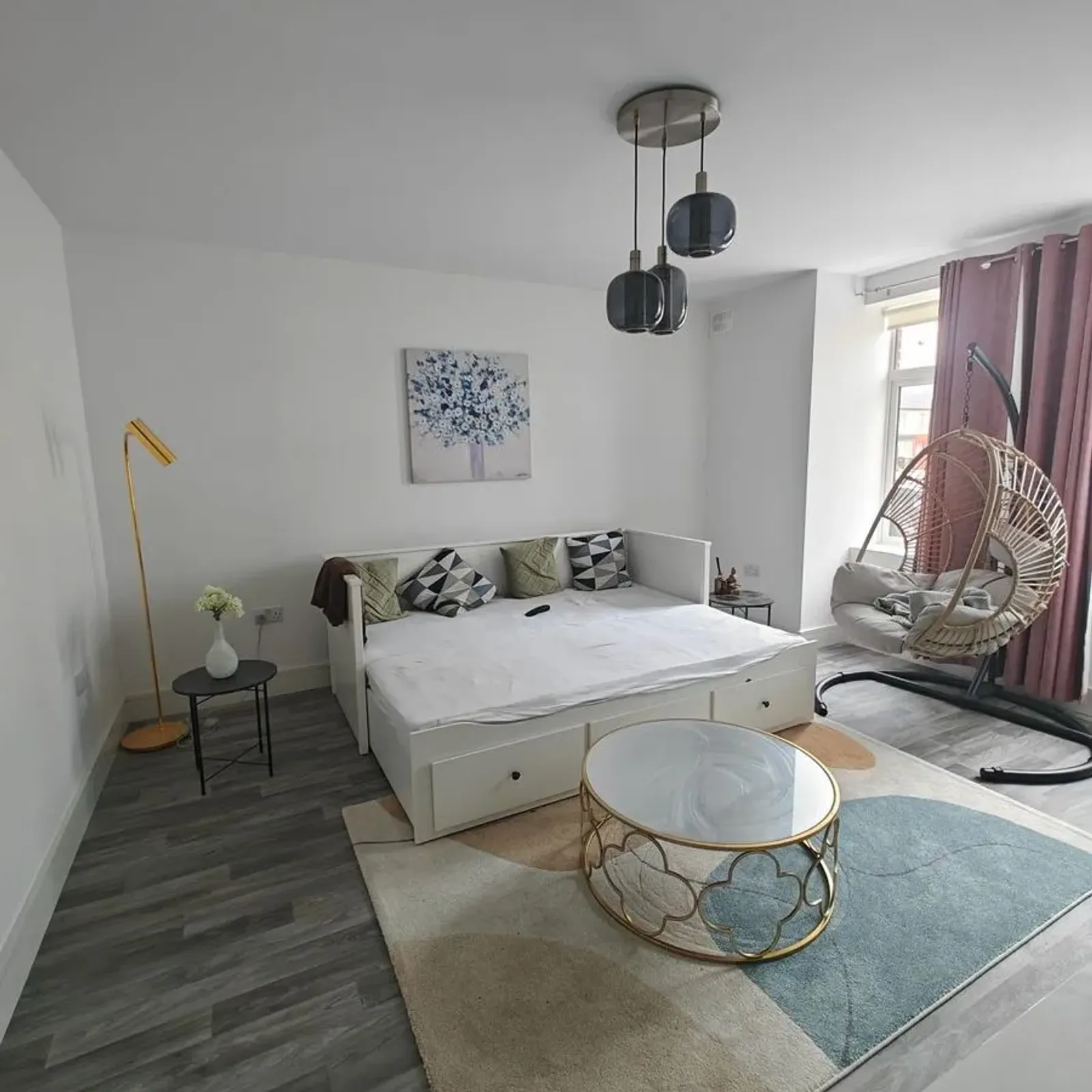 Bedroom with a white daybed, decorative pillows, round glass coffee table, hanging rattan chair by a window, pendant lights, and a floor lamp