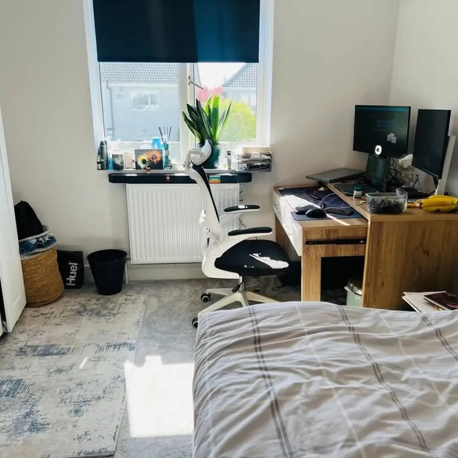 Bedroom with bed in foreground, desk with dual monitors and white office chair positioned by a window with a roller blind.