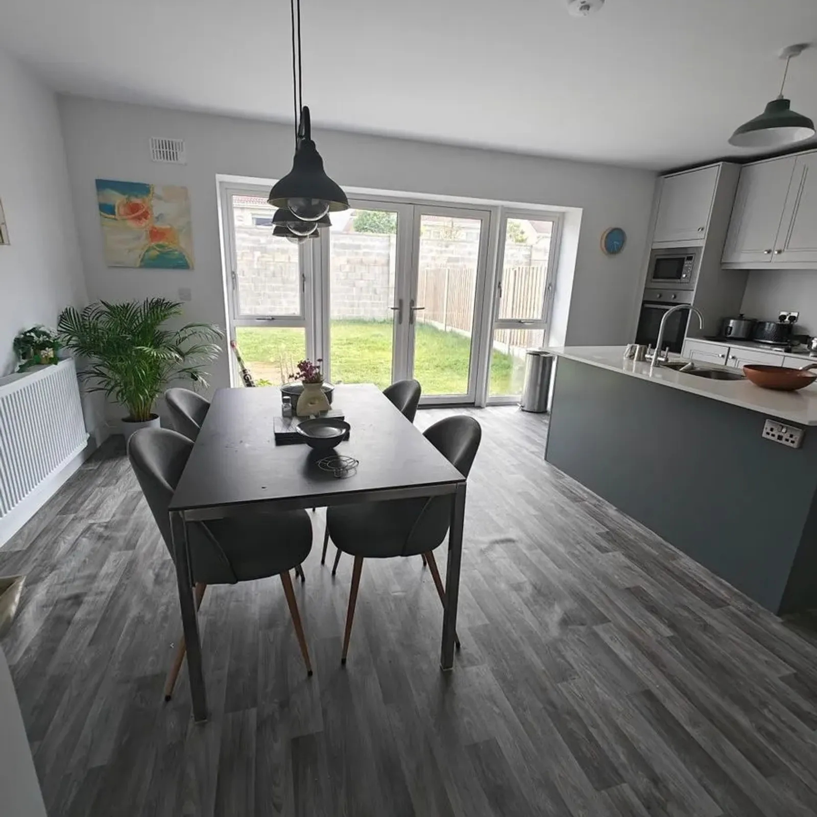 Open-plan dining area and kitchen with a rectangular dining table and four chairs, kitchen island with sink, pendant lights, and glass patio doors opening to a small lawn.