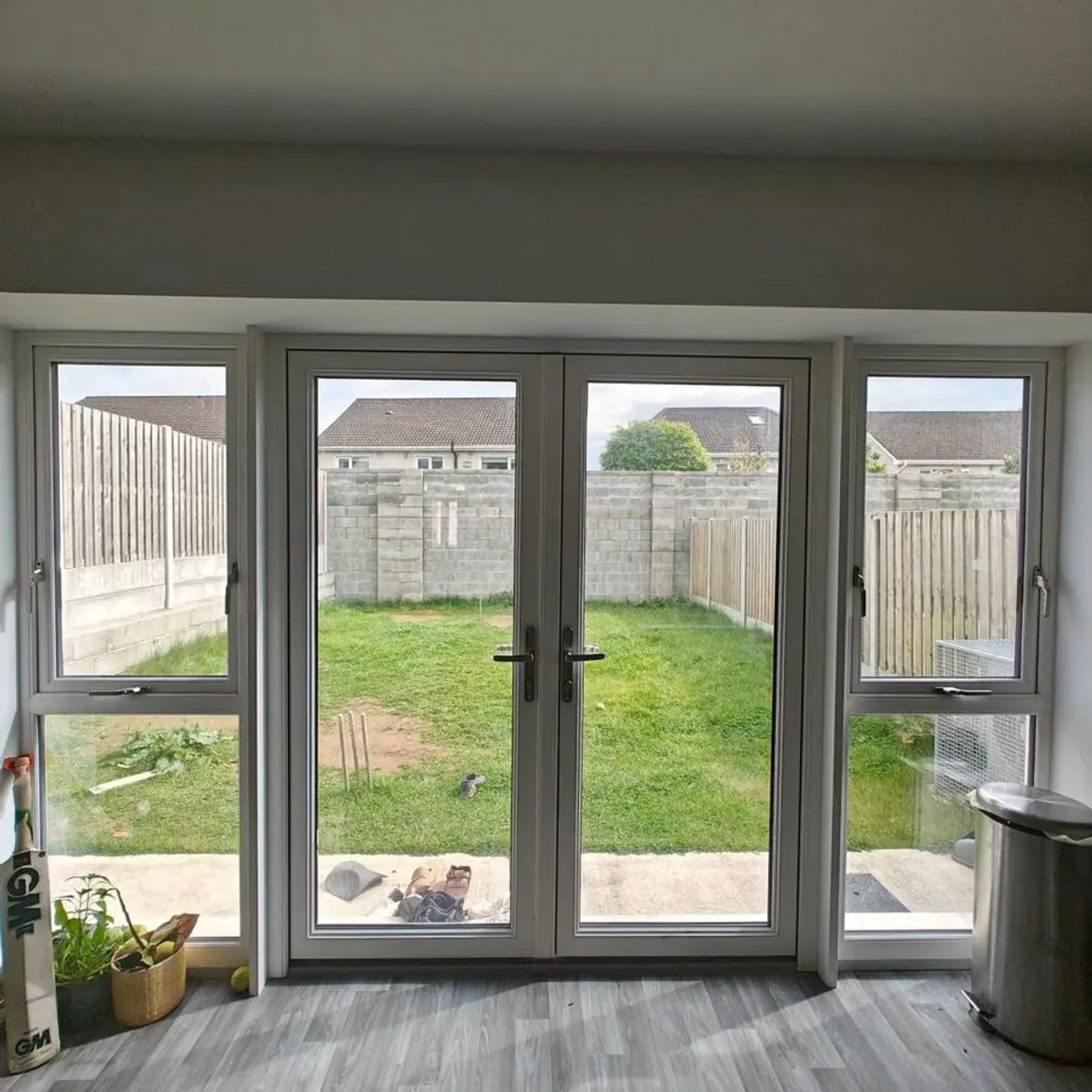Room with grey wood-look flooring and central glass double doors flanked by tall windows looking out onto a small grassy backyard and concrete patio. A plant is on the left windowsill and a trash bin is on the right.