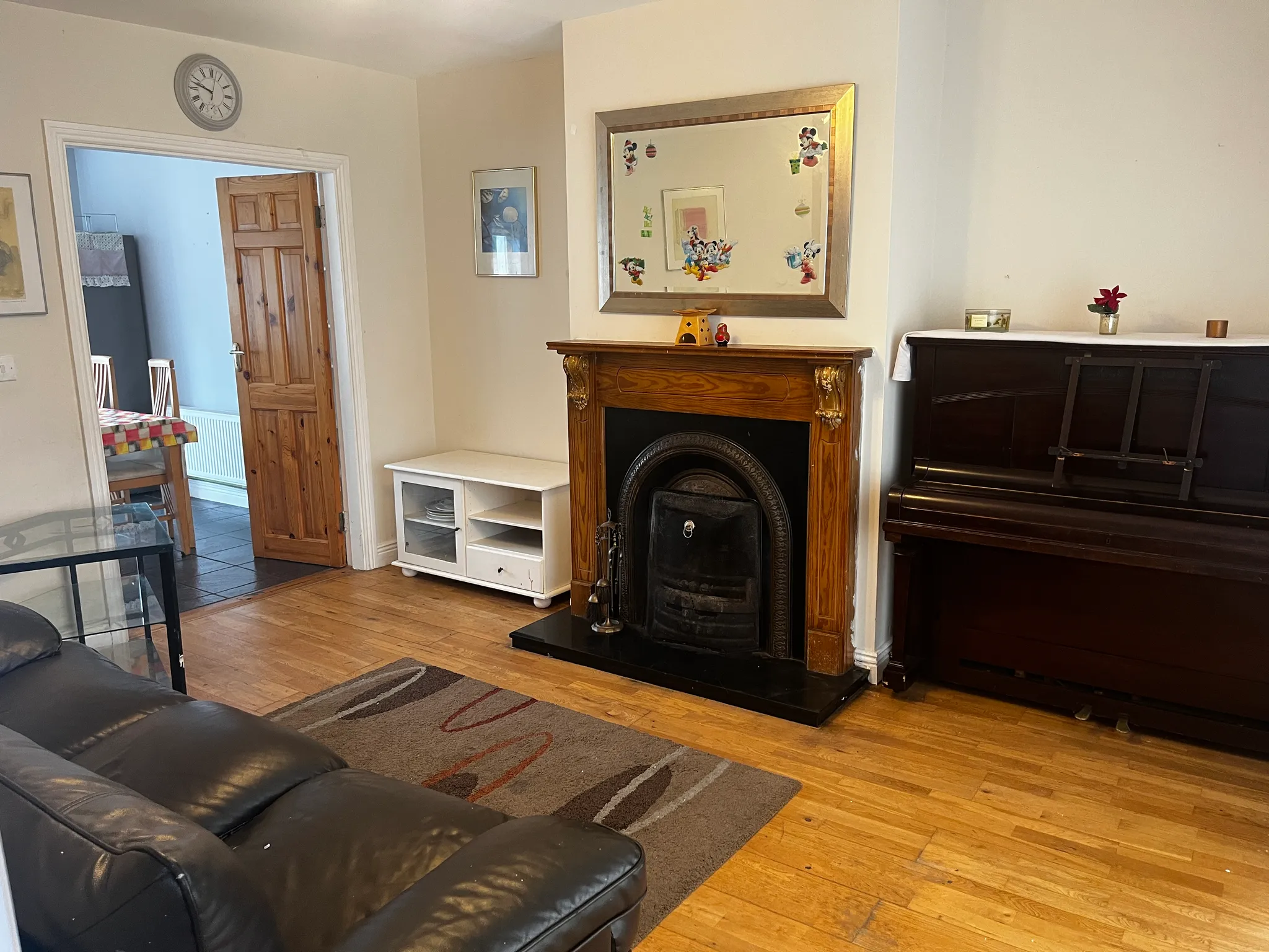 Living room with a fireplace and large framed mirror above it, upright piano, black leather sofa and a doorway leading to a dining area.