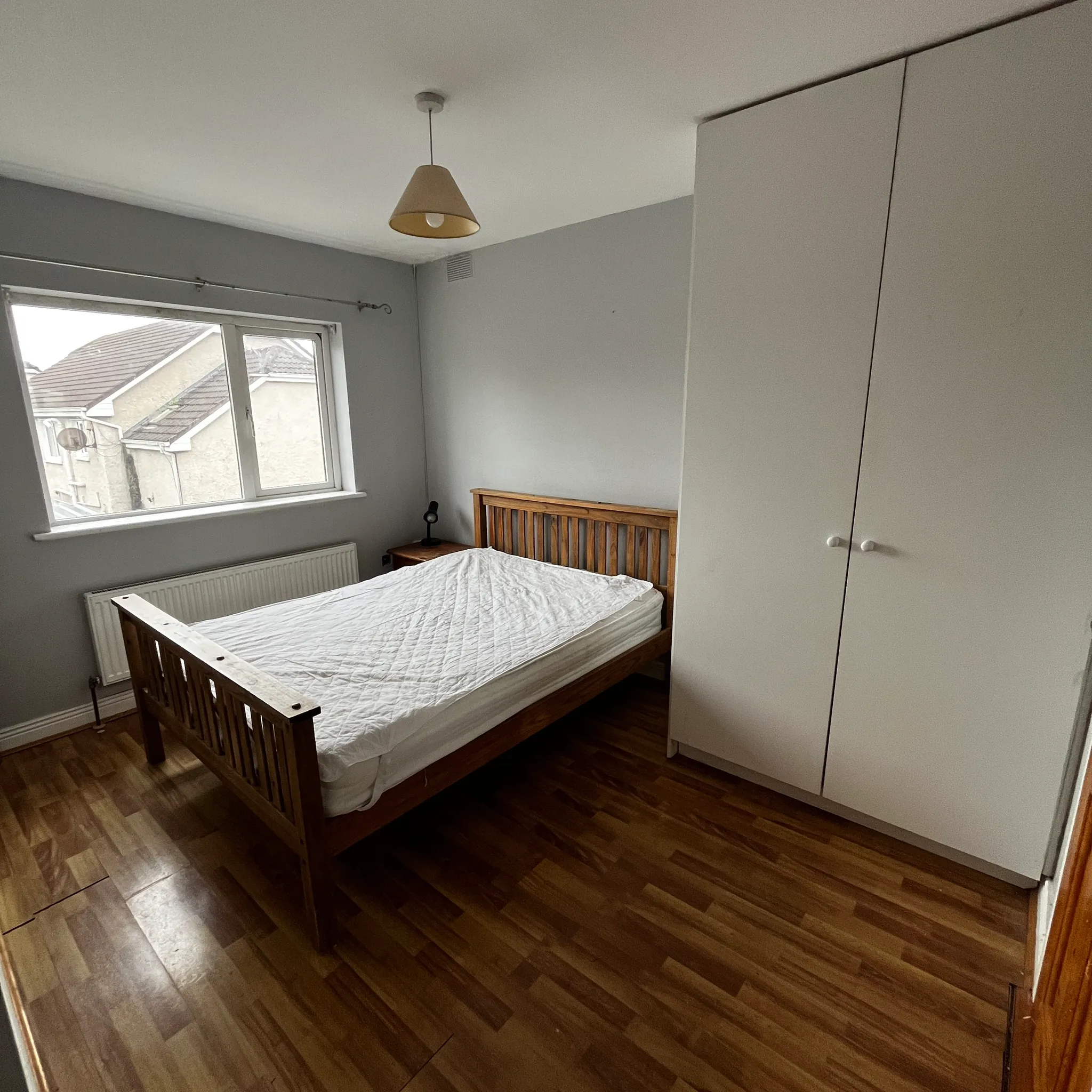 Bedroom with a wooden bed and mattress, white wardrobe, bedside table, radiator and a window with a view of neighbouring roofs.
