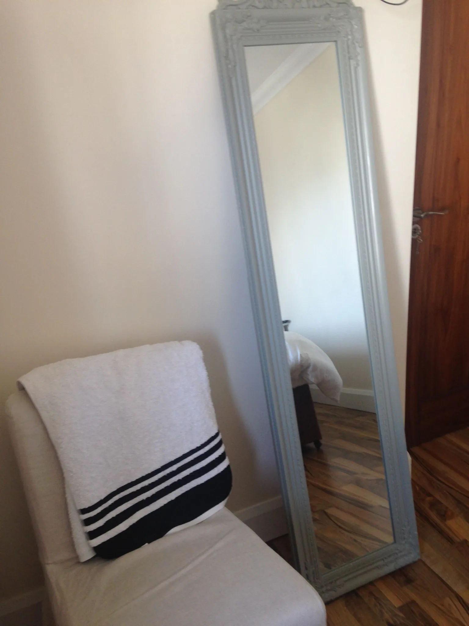 Corner of a bedroom showing a full-length framed mirror leaning against the wall, a chair with a folded towel, and a partially visible wooden door and bed reflected in the mirror.