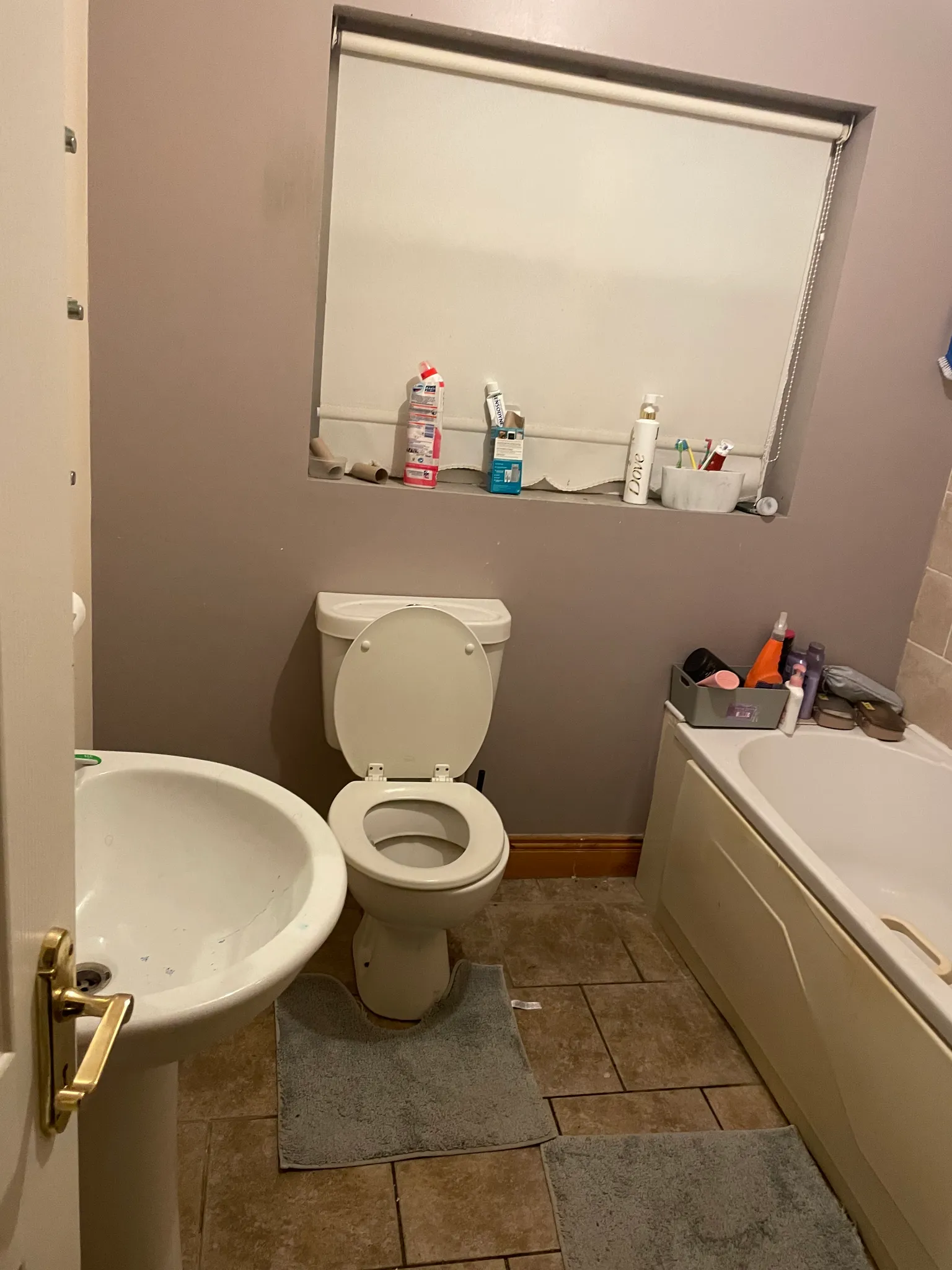 Bathroom with a toilet (seat up), pedestal sink, bathtub, small window with a roller blind, and toiletries on the windowsill and tub edge.