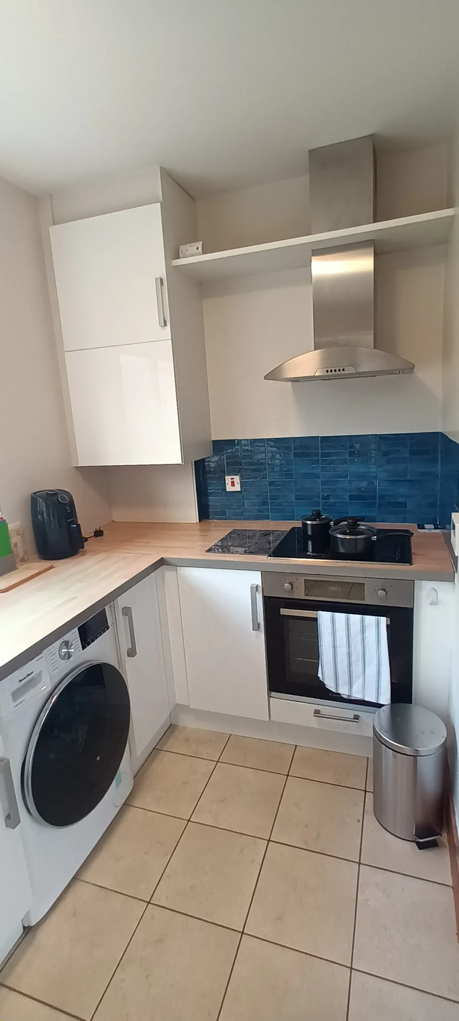 Kitchen with L-shaped countertop, electric cooktop and oven, stainless steel range hood, blue tile backsplash, white cabinets, washing machine and small trash bin