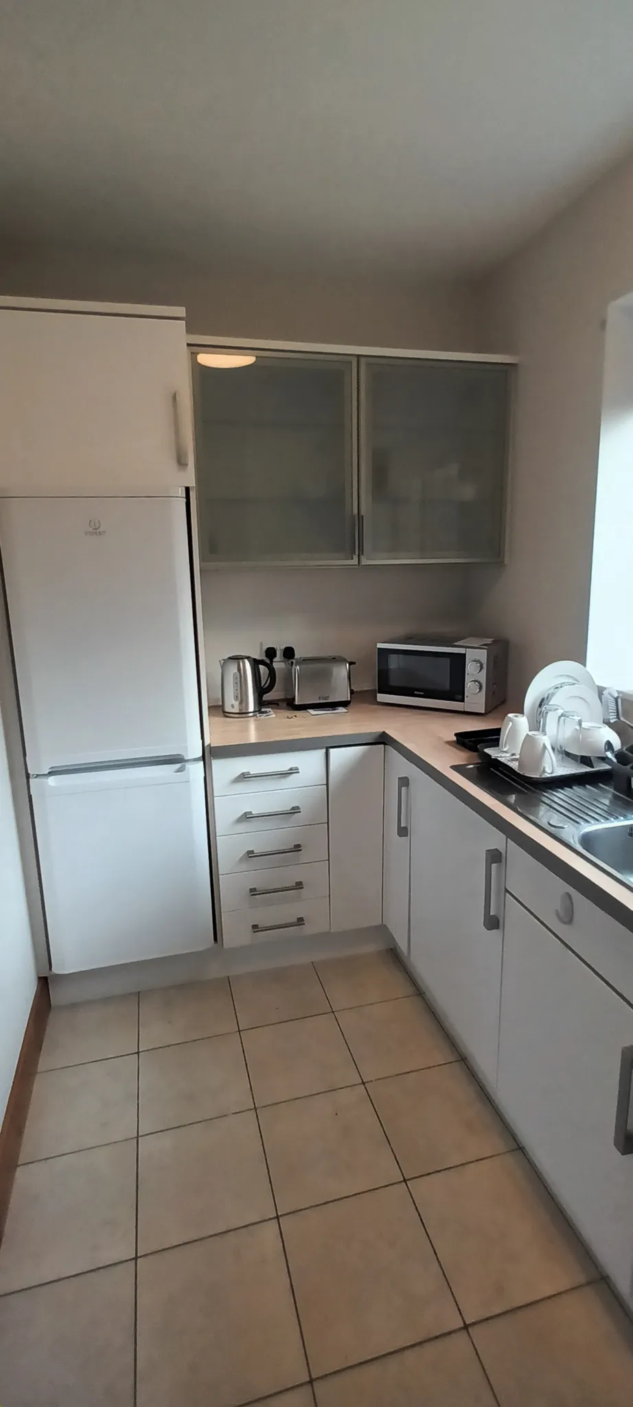 Kitchen with white cabinets, fridge, countertop appliances (kettle, toaster, microwave) and a sink with a dish rack