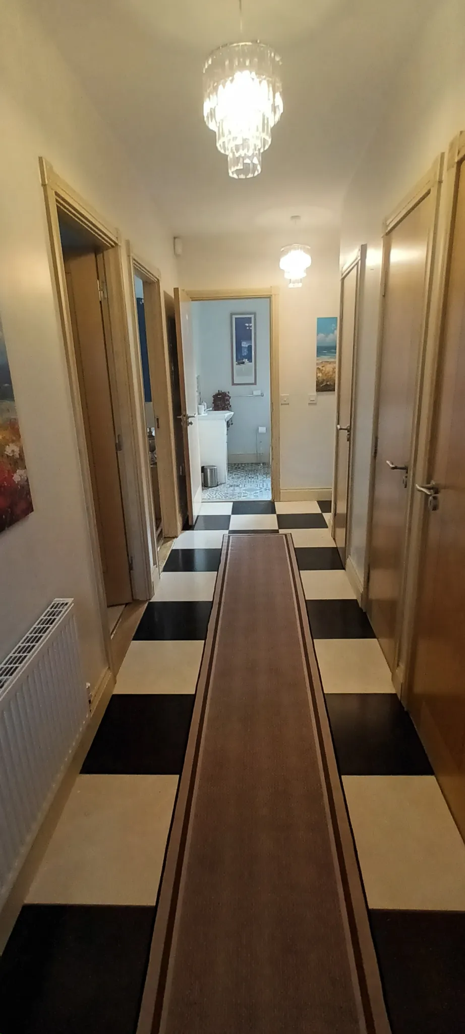 Hallway with checkerboard floor, long runner rug, multiple closed doors on both sides and ceiling lights; a bathroom doorway is visible at the far end.