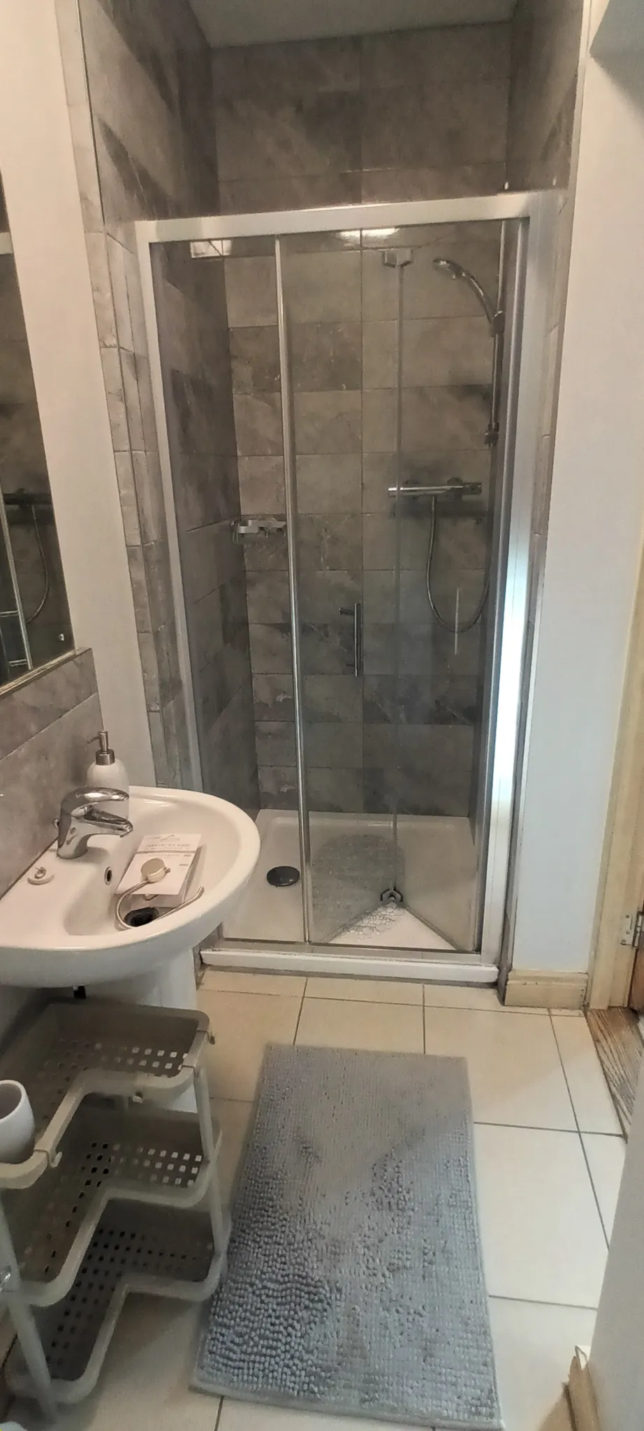 Bathroom with an enclosed tiled shower with glass sliding door, a pedestal sink with soap dispenser and small shelving unit, and a bath mat on tiled floor.
