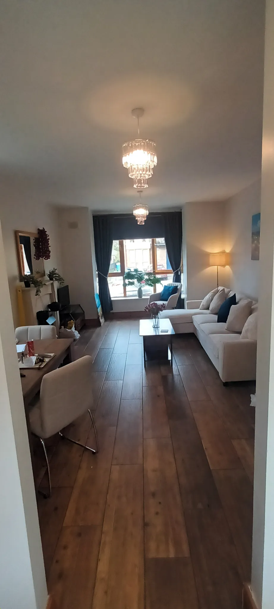 Living room with a beige sectional sofa, coffee table, wooden floor, window with dark curtains, pendant lights, and a dining table at the left