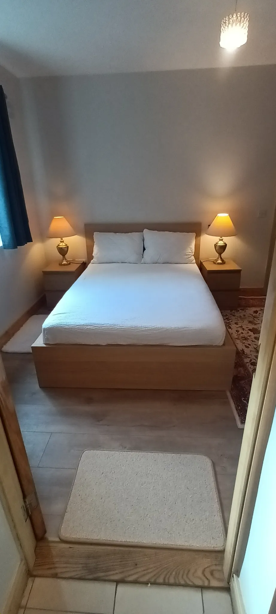 Bedroom with a double bed centered between two bedside tables each with a lamp, a window with a dark curtain at left, and rugs on the floor.