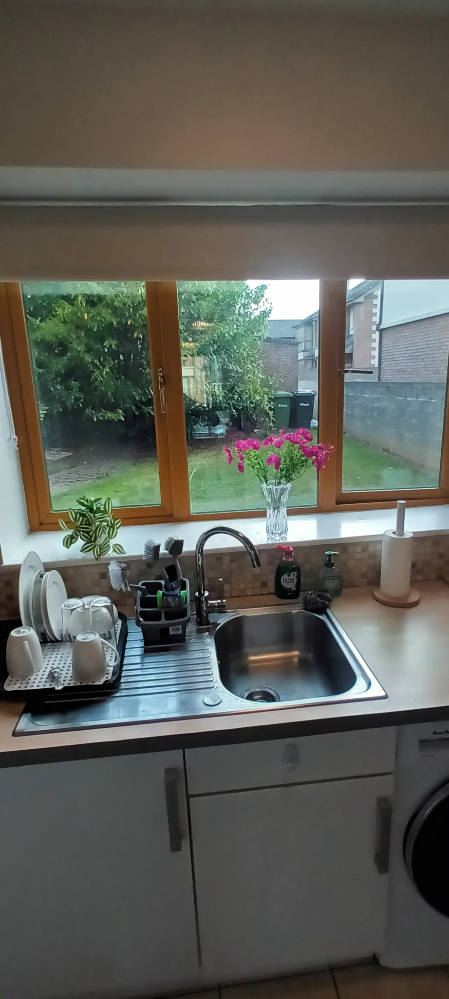 Kitchen sink and countertop beneath a three-panel window with a garden view; vase of flowers and dish rack on the windowsill and worktop.