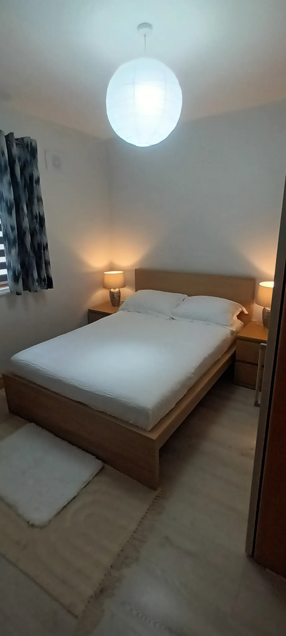 Bedroom with a double bed, wooden headboard, bedside tables with lamps, pendant ceiling light and a window with patterned curtains.