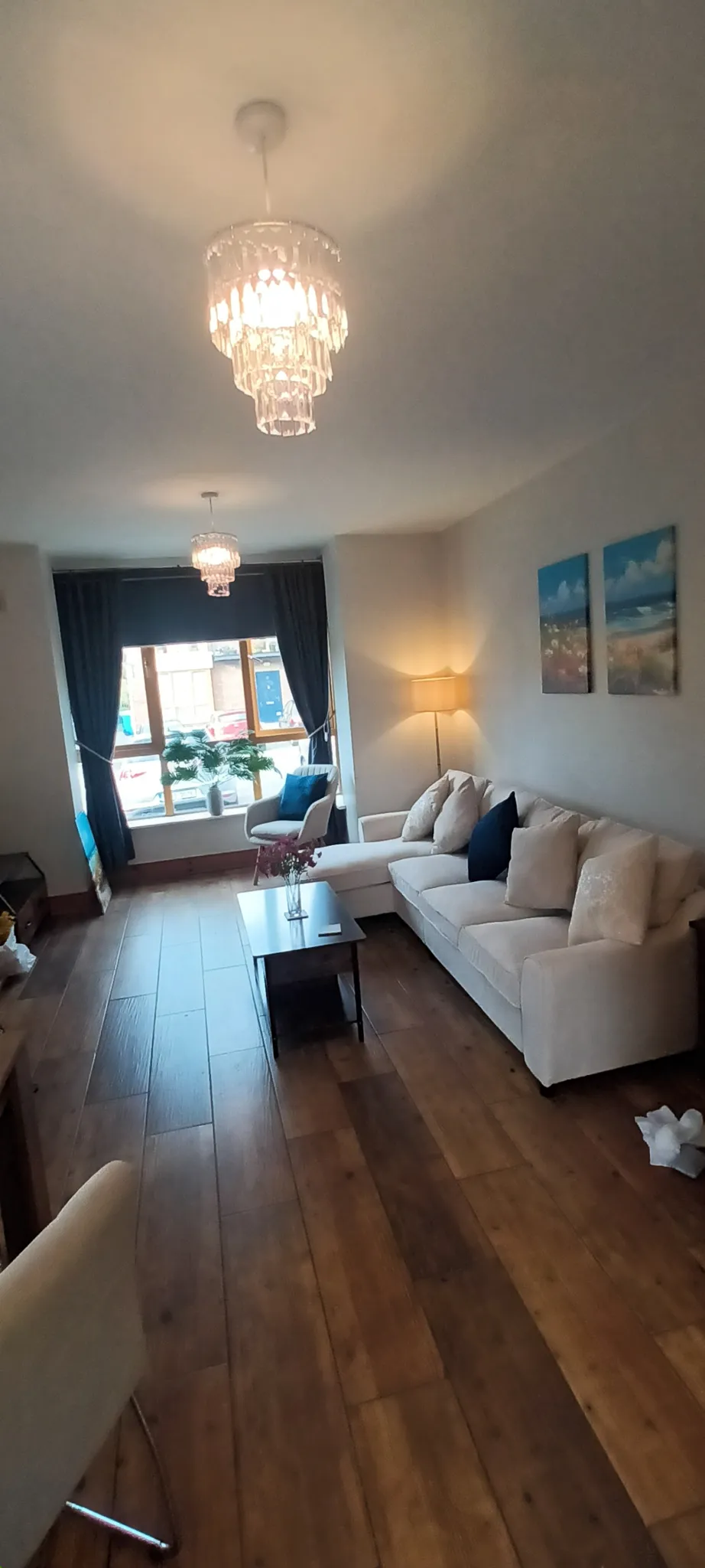 Living room with a beige sectional sofa and cushions, coffee table on a wood floor, armchair by a window with dark curtains, two hanging chandeliers and a floor lamp, wall art above the sofa.