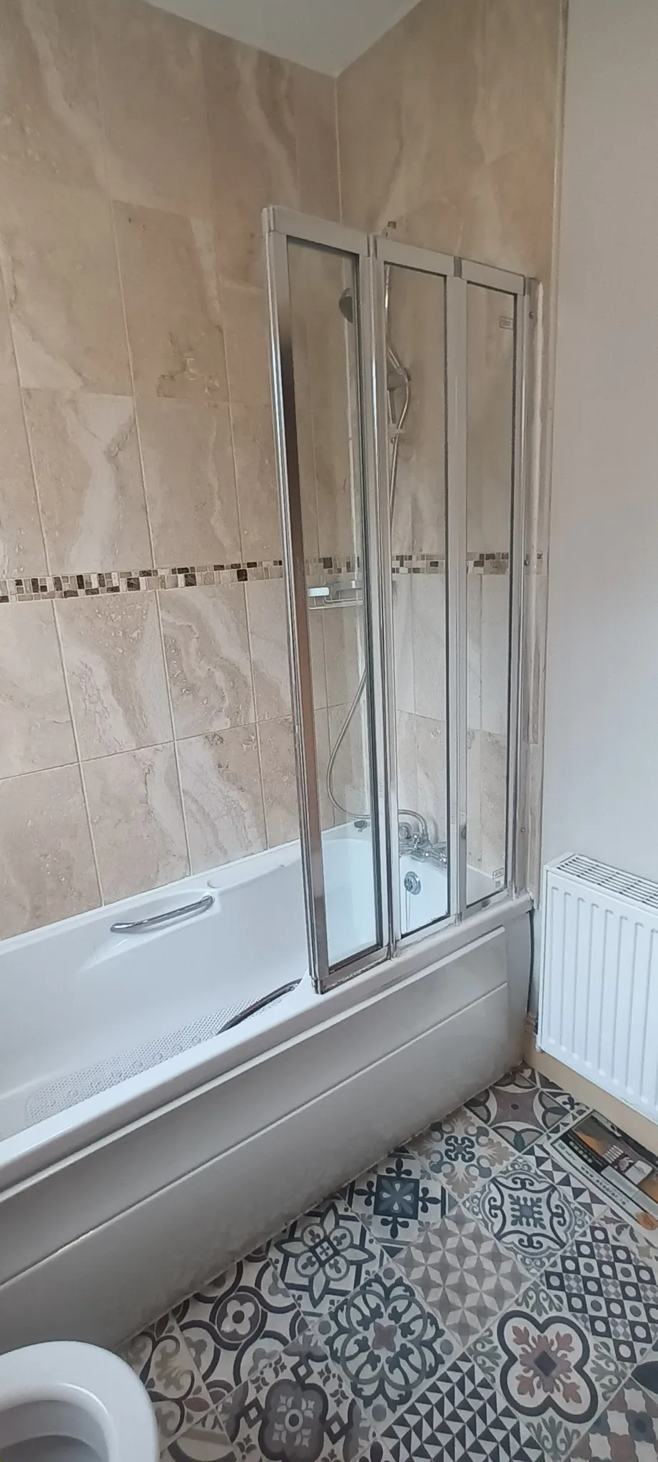 Bathroom with a bathtub and folding glass shower screen, wall tiles with mosaic border and patterned floor tiles, radiator visible.