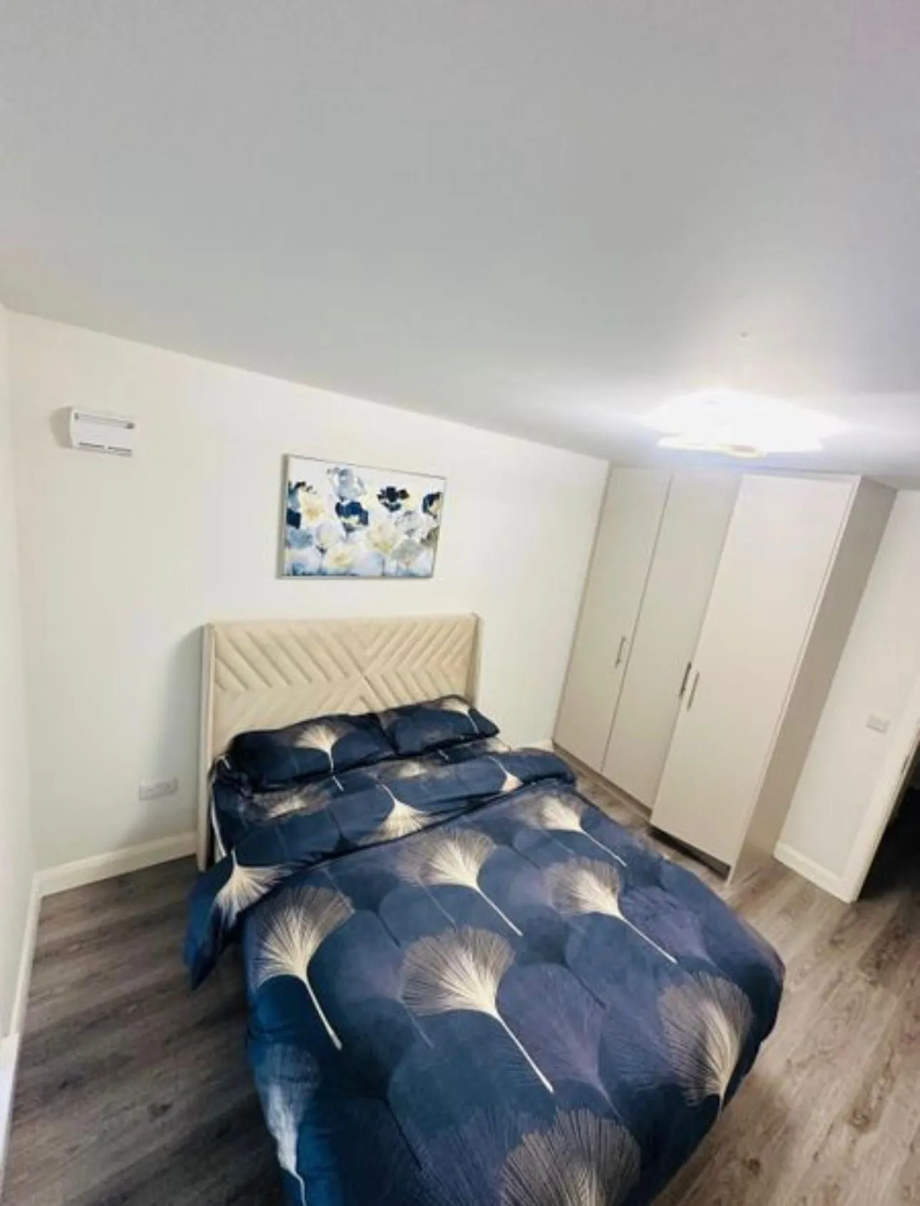 Bedroom with a double bed with blue patterned duvet, beige upholstered headboard, white wardrobe, wall art above the bed and ceiling light