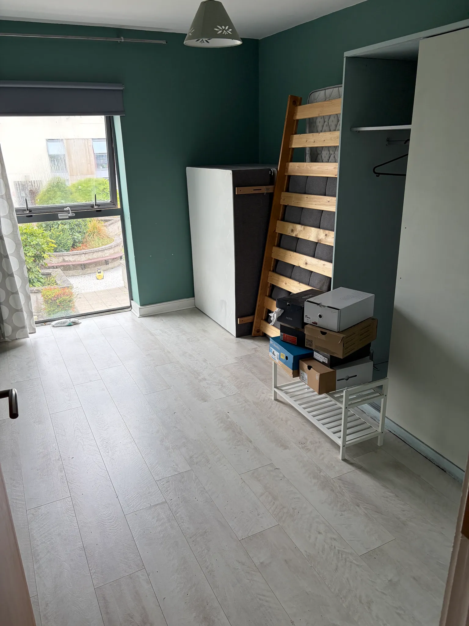 Bedroom with a stacked mattress and wooden bed slats leaning against a teal wall, open closet with hangers, a window with a roller blind on the left, and a small rack with stacked shoe boxes on the floor.