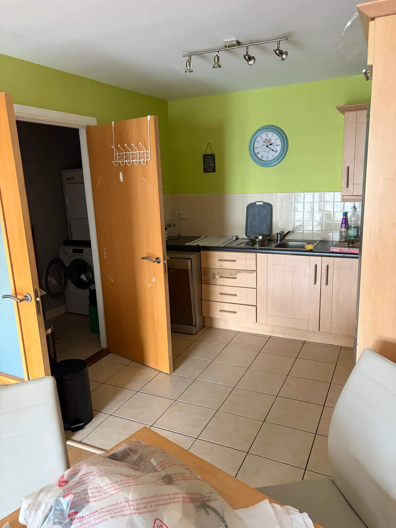 Kitchen with light wood cabinets, tiled backsplash and floor, a sink and countertop, open wooden door showing stacked washer and dryer in a dark adjoining room, and a wall clock on a green wall.