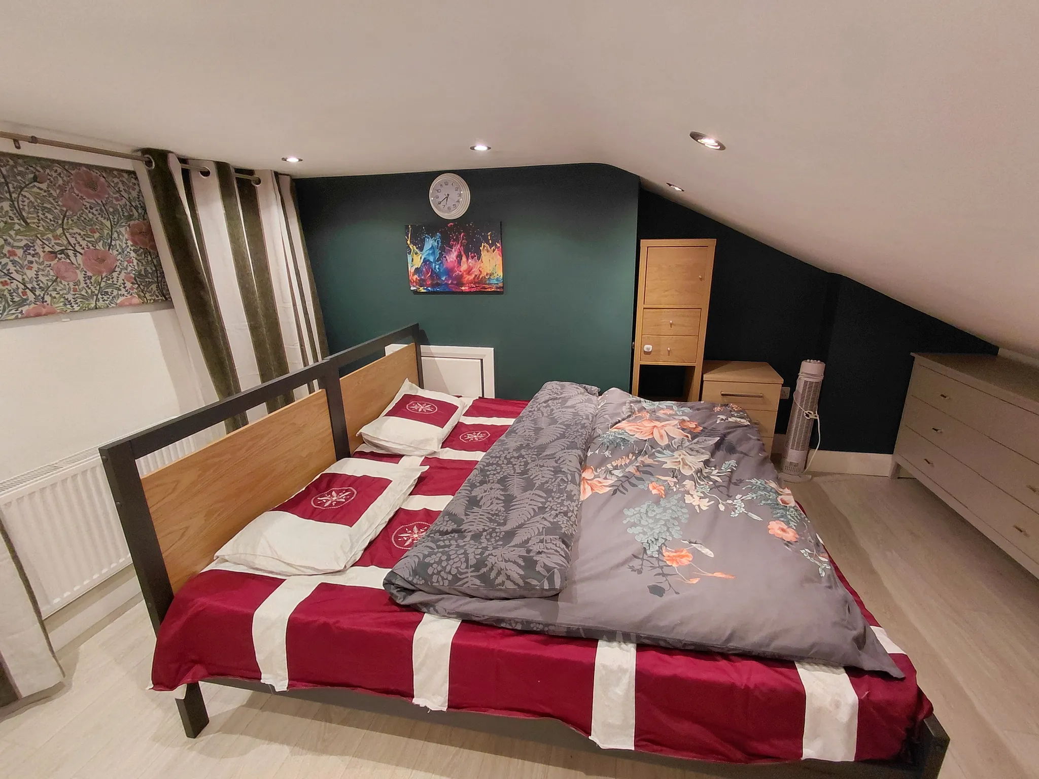 Bedroom with a double bed and patterned bedding, sloped ceiling, curtains, dresser and a small chest of drawers with wall art and clock above the bed.