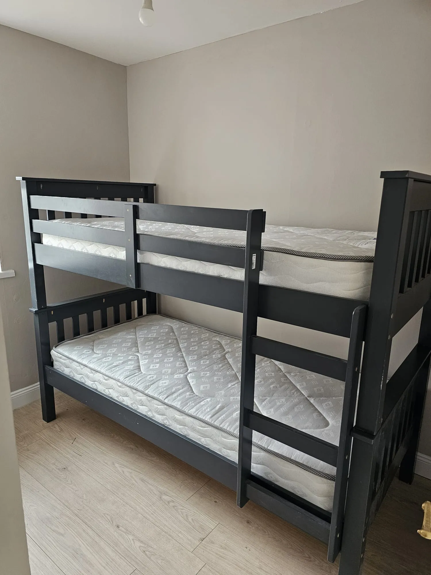 Bedroom with a dark wooden bunk bed, two mattresses and a ladder against a plain light-colored wall.