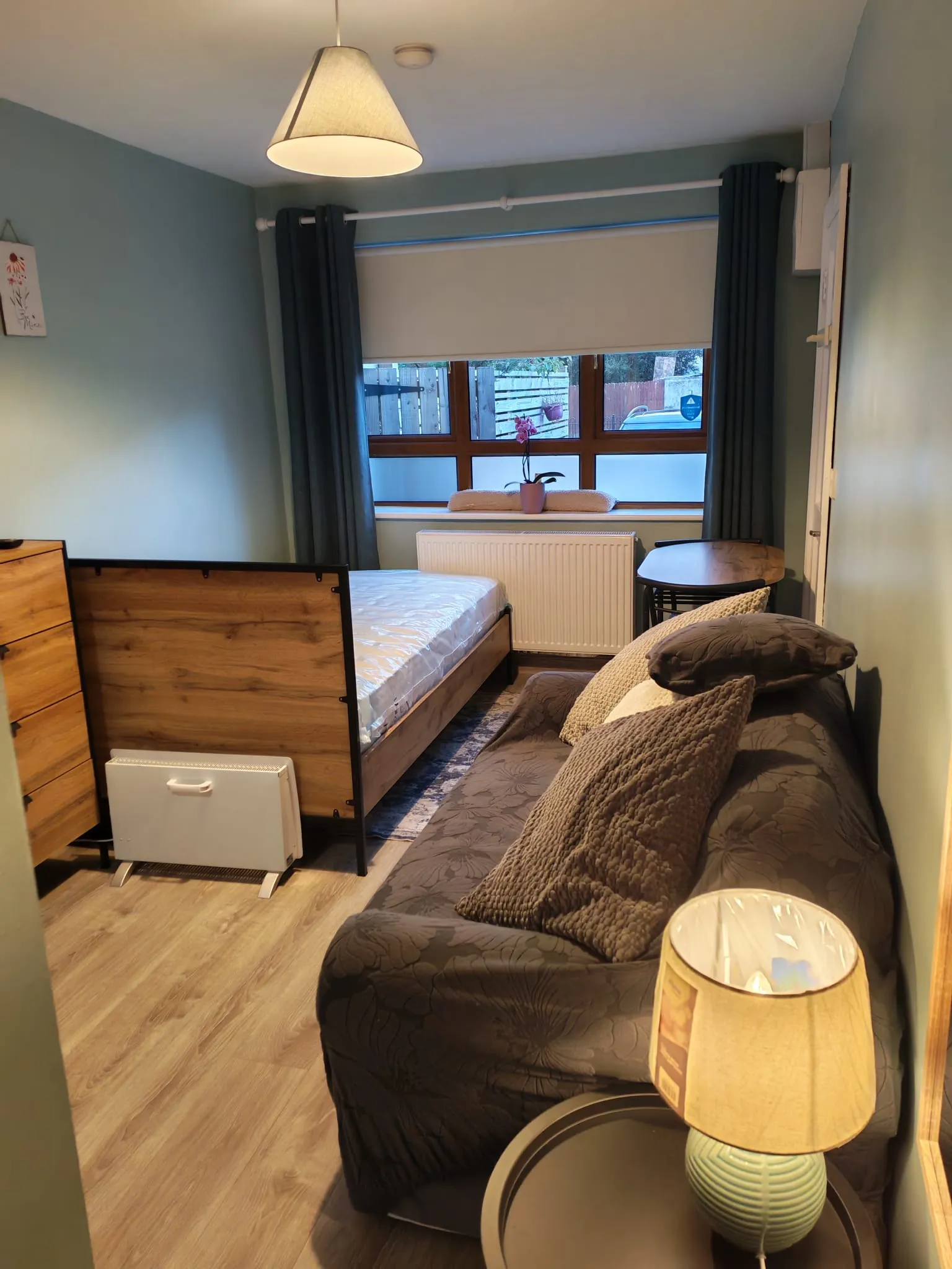 Bedroom with a single bed frame, sofa with cushions, window with roller blind and curtains, radiator and bedside lamp