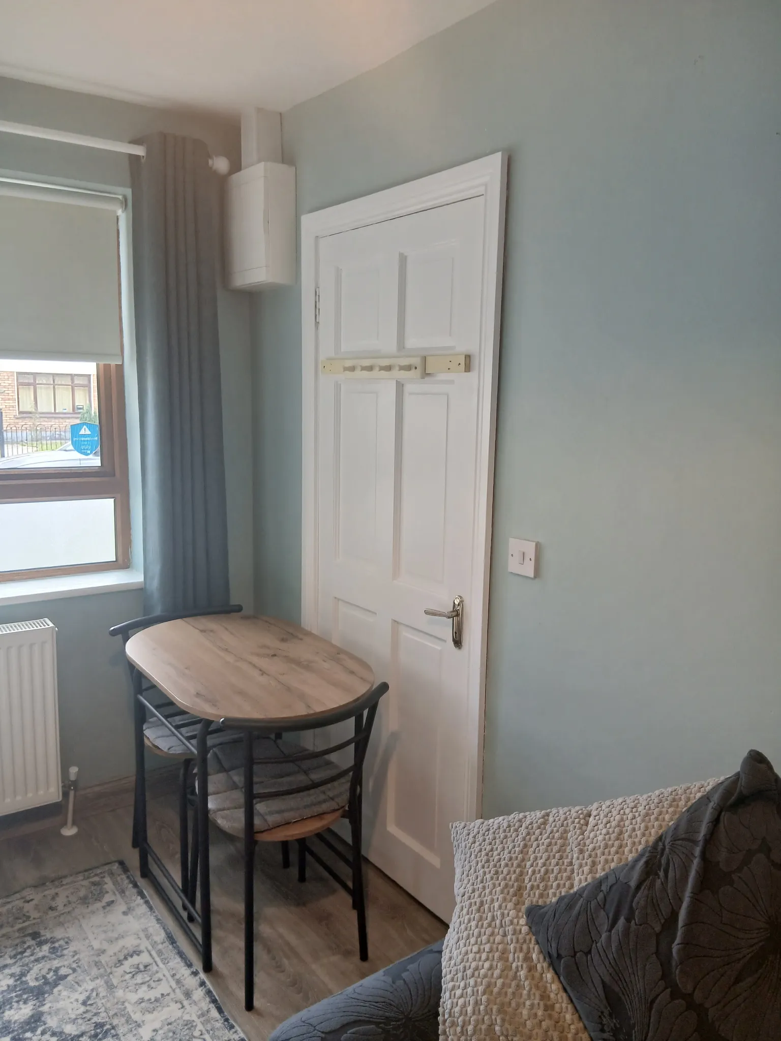 Corner of a living room with a small wooden table and two chairs next to a closed white door and a window with a roller blind and curtain.