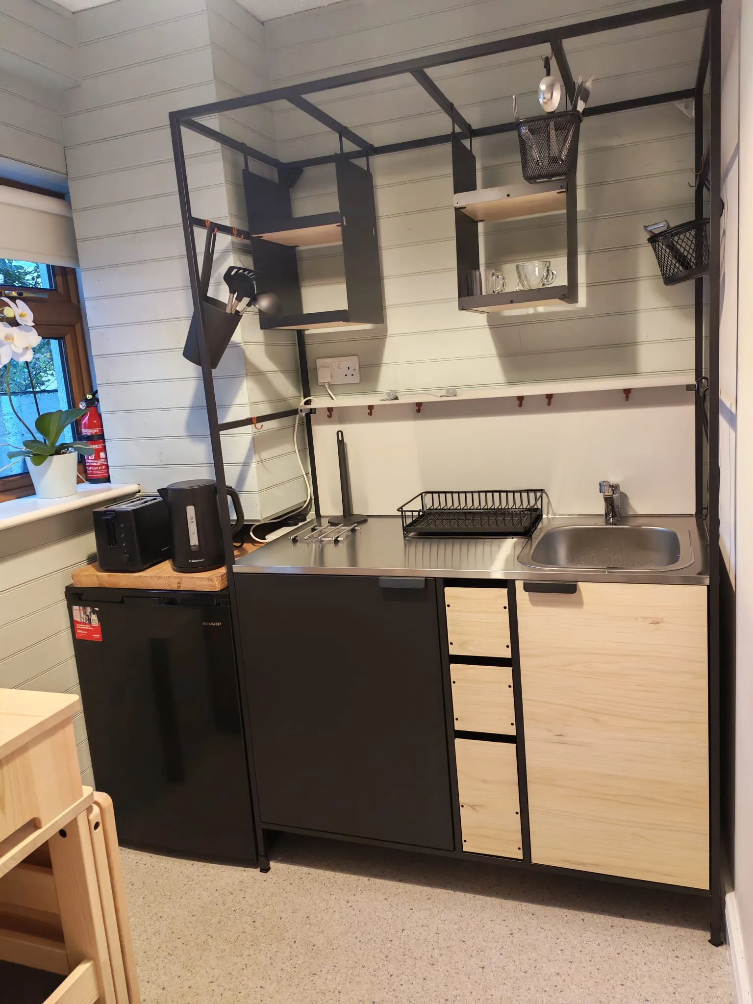 Small kitchen area with a stainless steel sink and countertop, open metal shelving above, dish rack, kettle and toaster, and a small refrigerator.