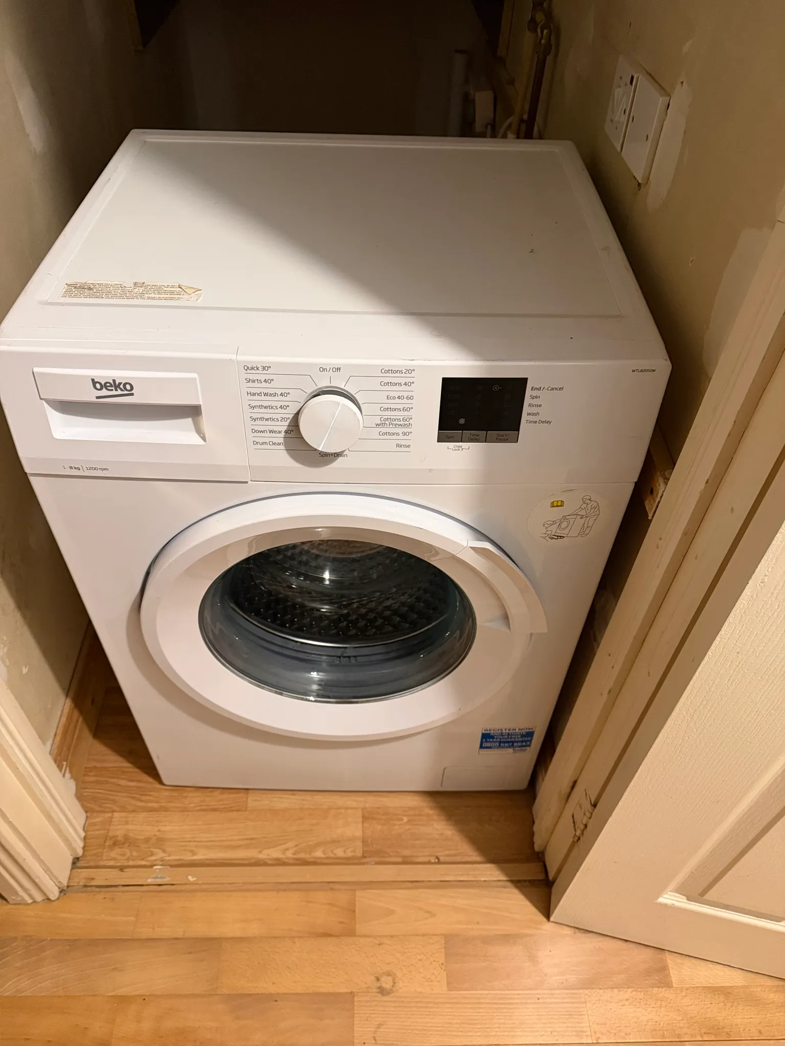 Front-loading washing machine in a narrow alcove with a wooden floor and a partially open door nearby.