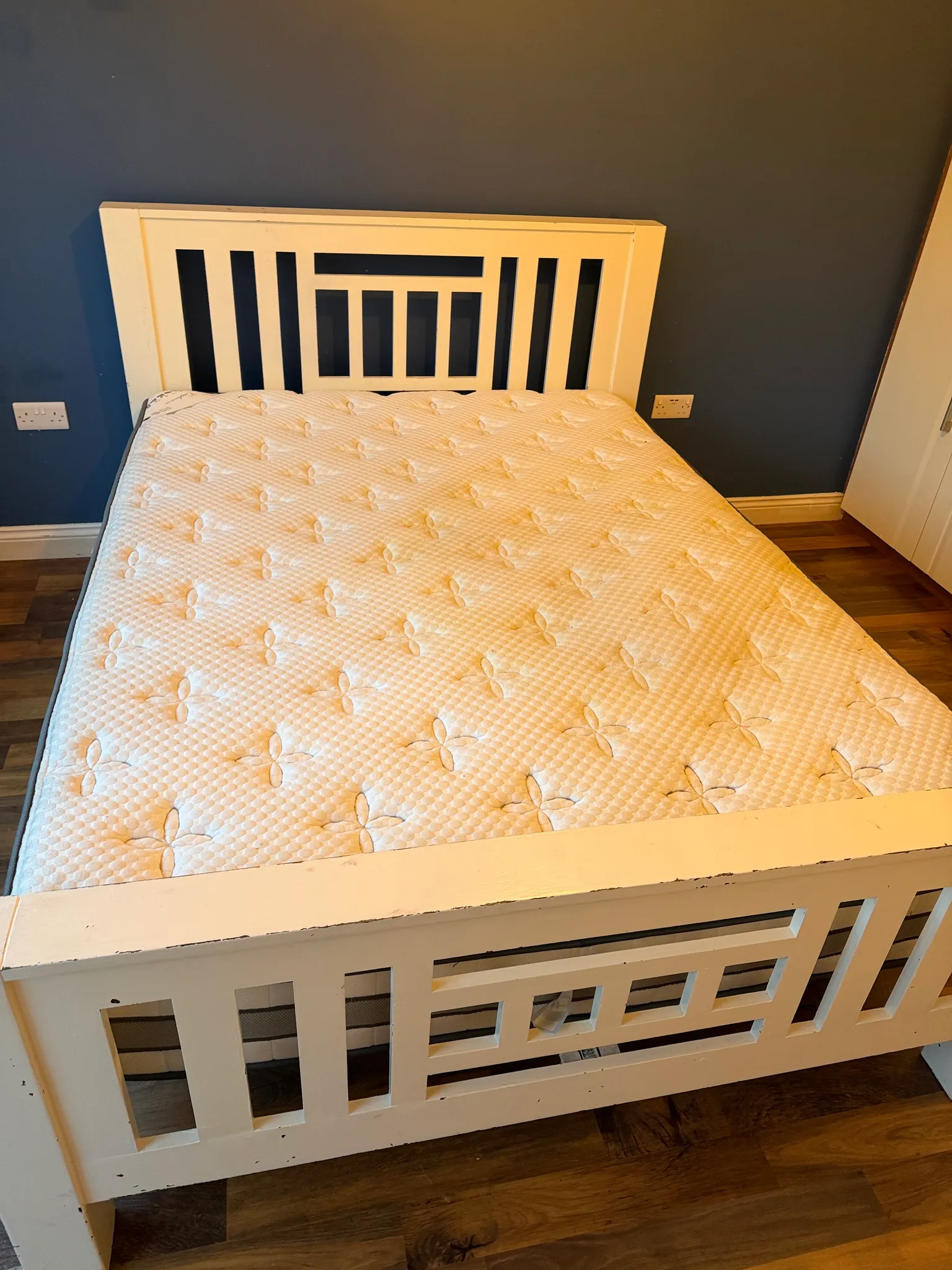 Bedroom corner showing a mattress on a white wooden bed frame with a slatted headboard against a dark blue wall