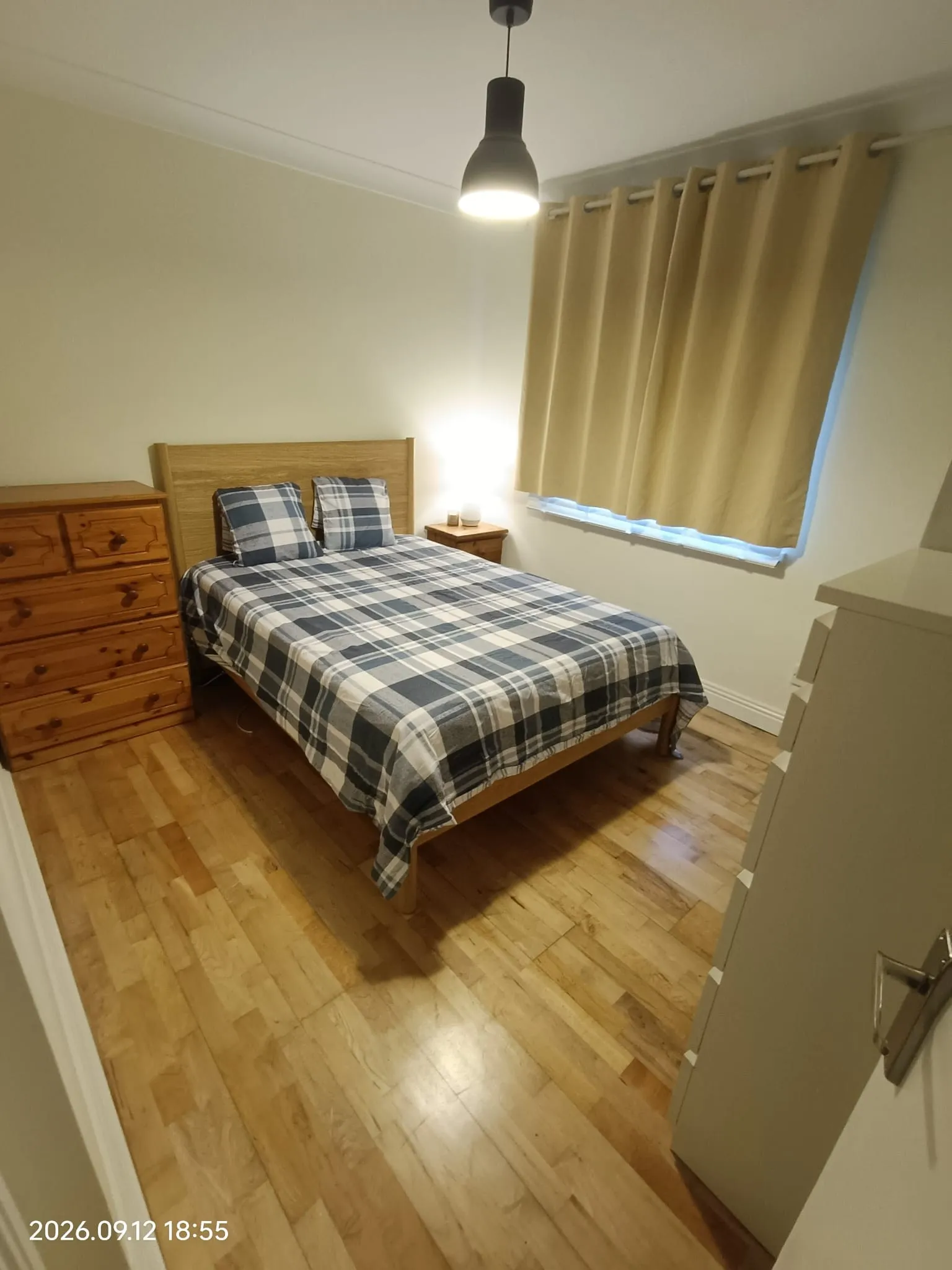 Bedroom with a double bed dressed in plaid bedding, wooden headboard, nightstand with a lit lamp, chest of drawers, pendant ceiling light and a curtained window