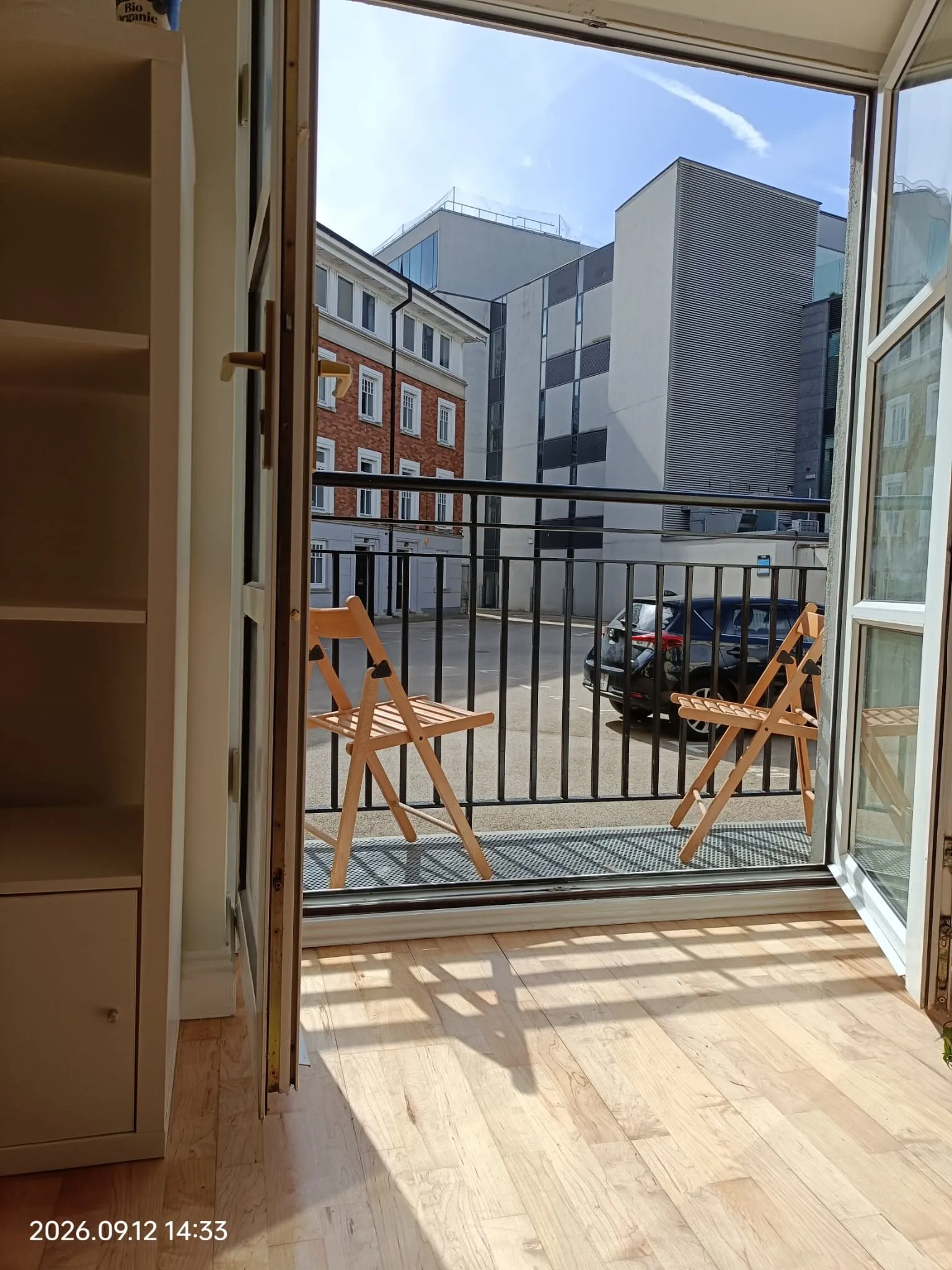Open window leading to a small balcony with two wooden folding chairs and a metal railing, view of neighboring buildings and a parked car.