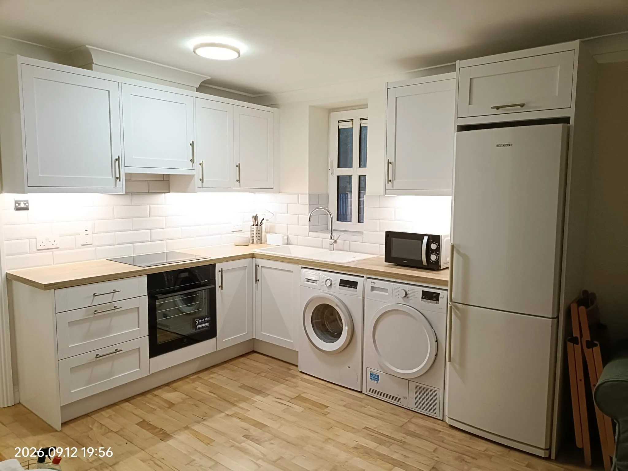 Kitchen with white cabinets and tiled backsplash, countertop with sink and faucet under a small window, refrigerator and microwave, oven and stacked washer and dryer.