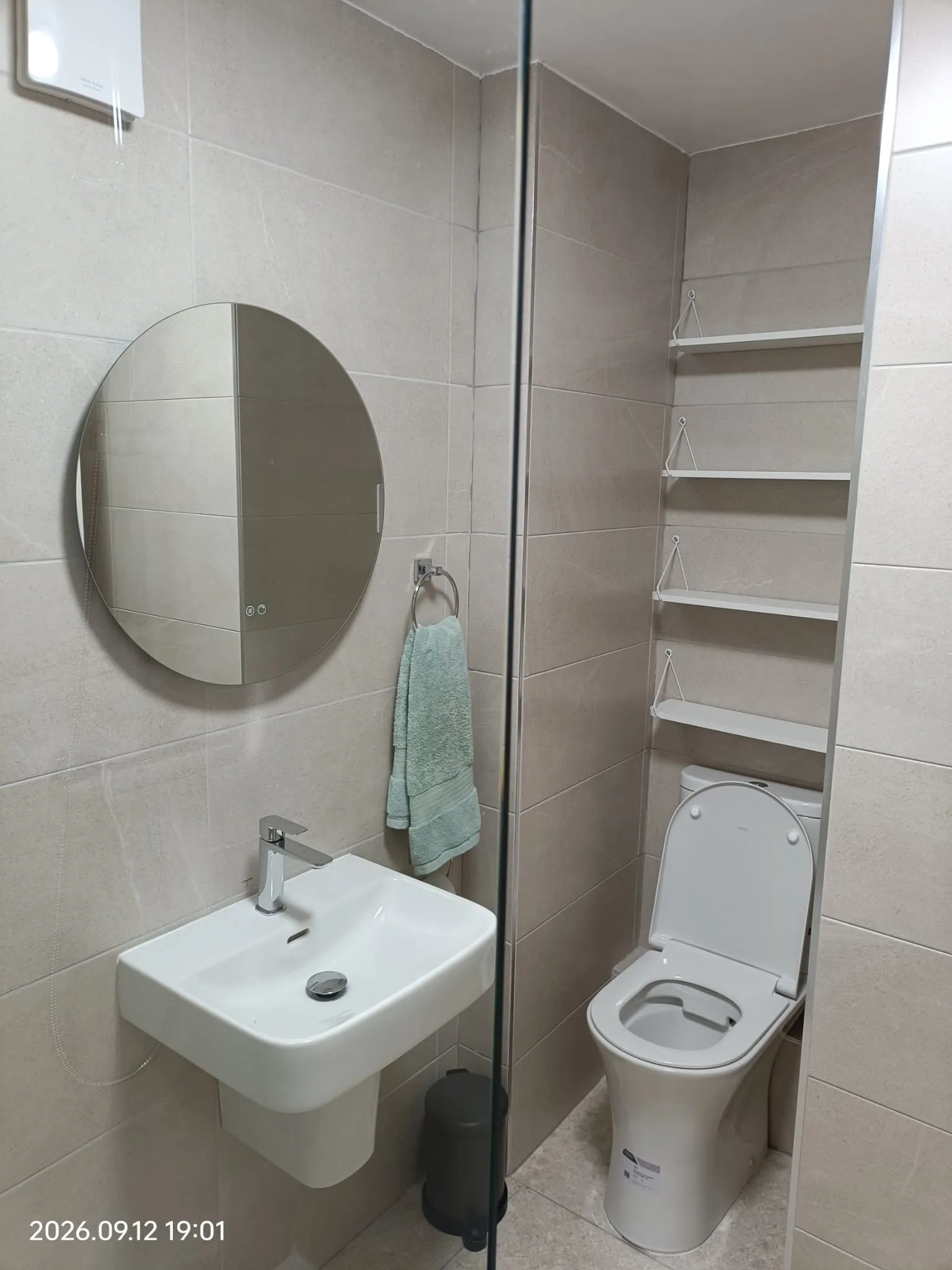 Bathroom with a wall-mounted sink and faucet, round mirror, toilet with open lid, towel on a ring, wall-mounted shelves, and a glass shower divider.