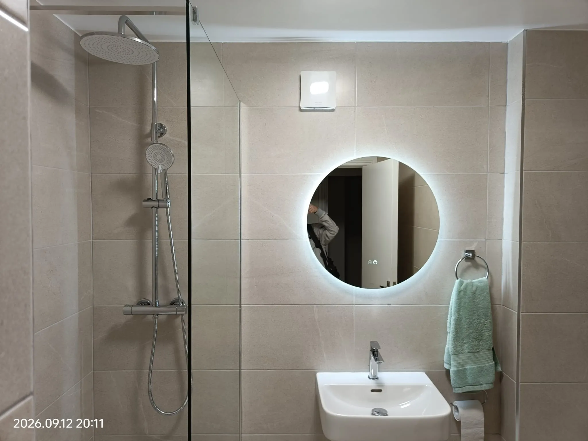 Bathroom with a wall-mounted sink, round backlit mirror, towel ring with green towel, and a shower with a glass screen and rainfall showerhead