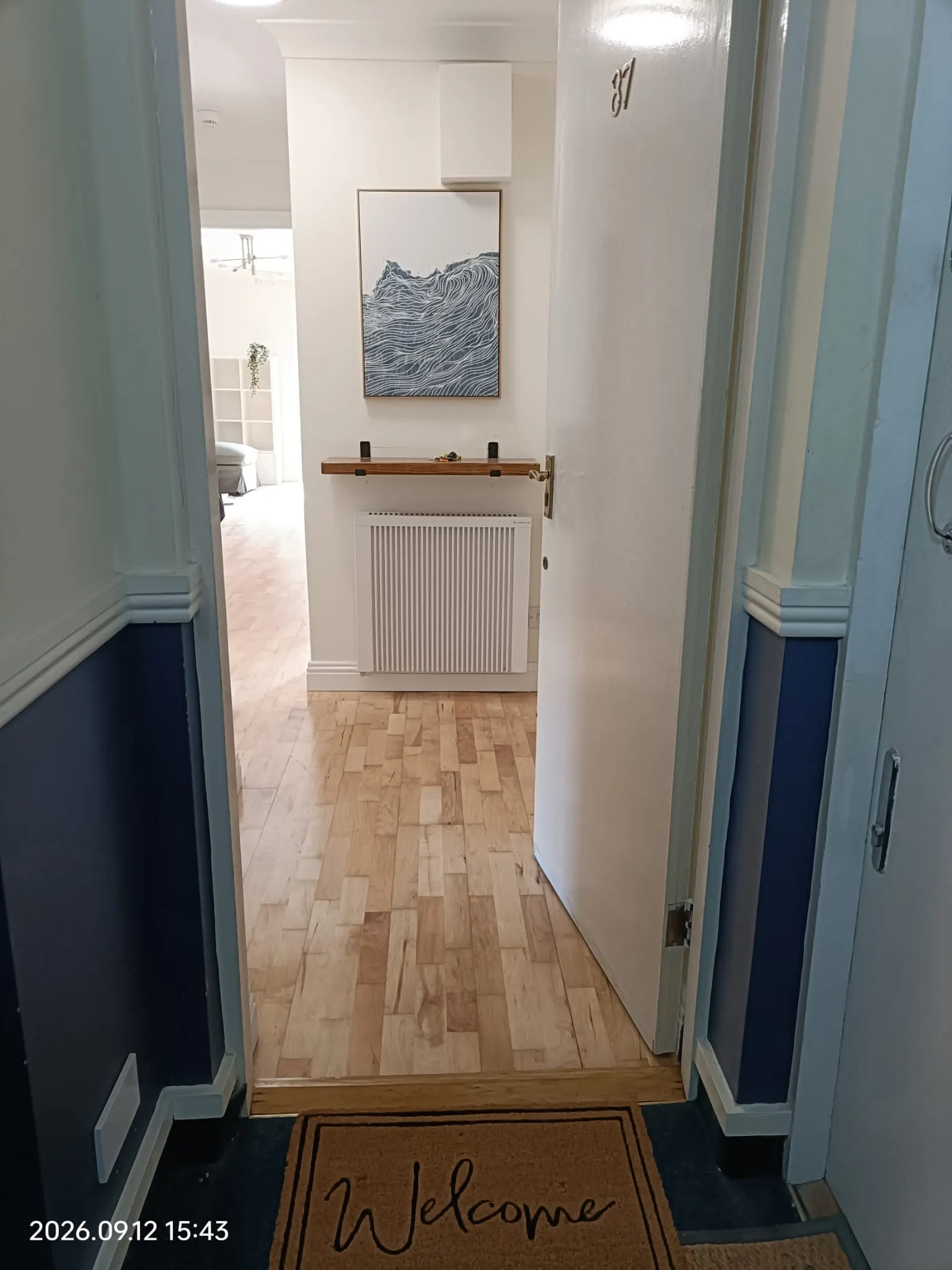View through an open entrance door into a hallway with wooden floor, radiator, small shelf and framed wall art; a welcome doormat is visible at the foreground.