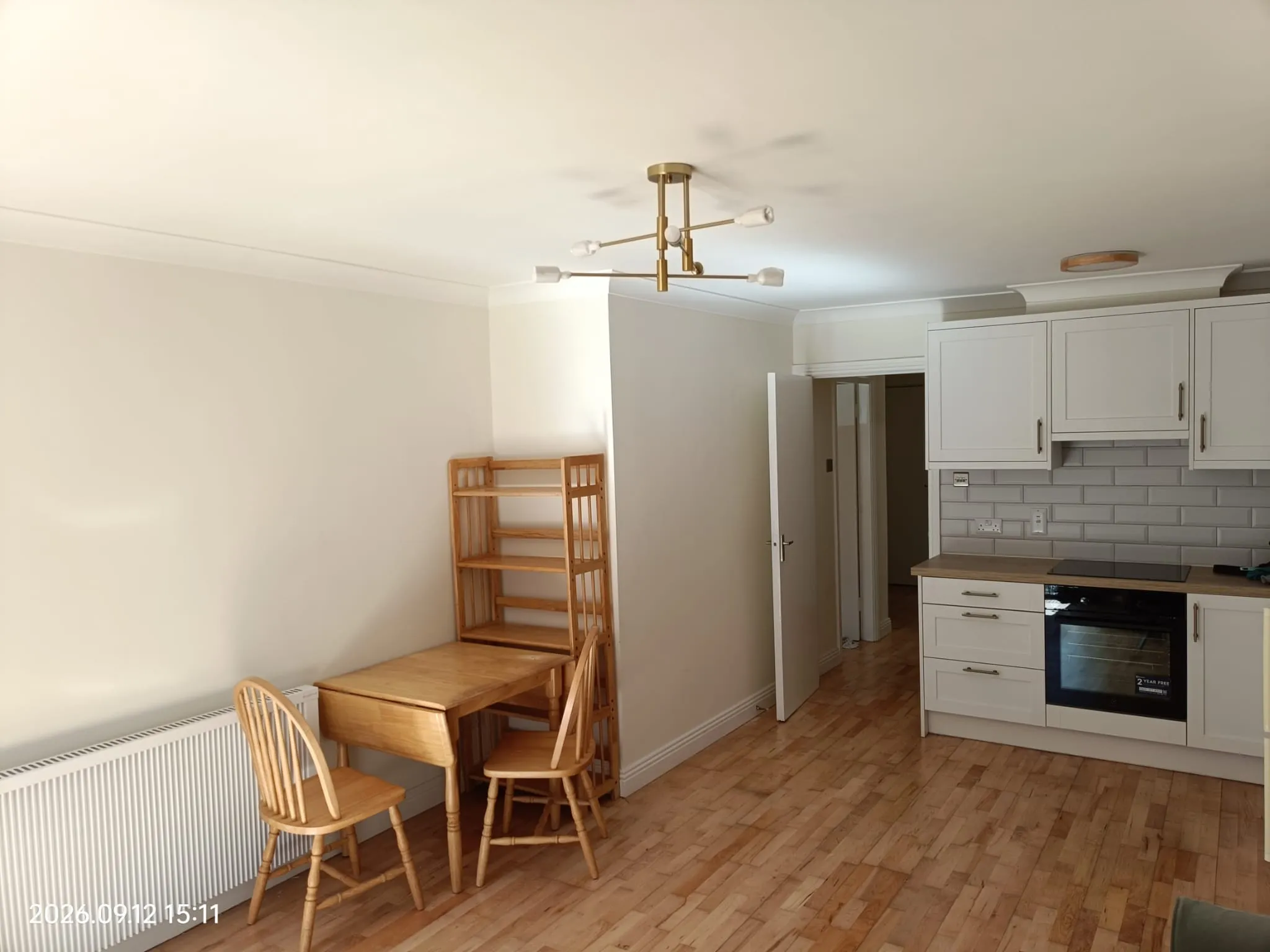Dining area and kitchen with a small wooden table and chairs, wooden shelving, white cabinets, built-in oven, tiled backsplash and ceiling light fixture.