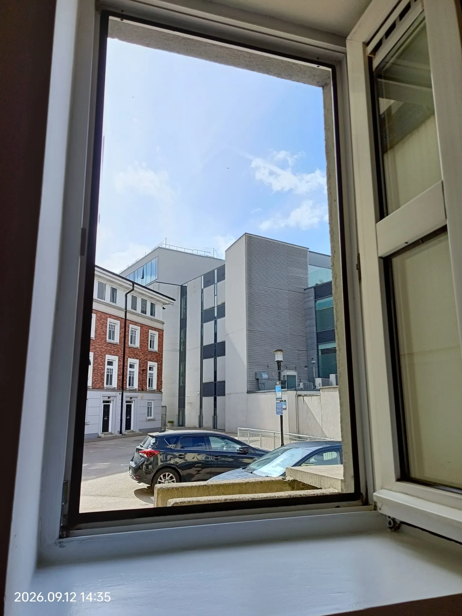 View through an open window showing a street with parked cars and nearby modern and brick buildings under a blue sky.