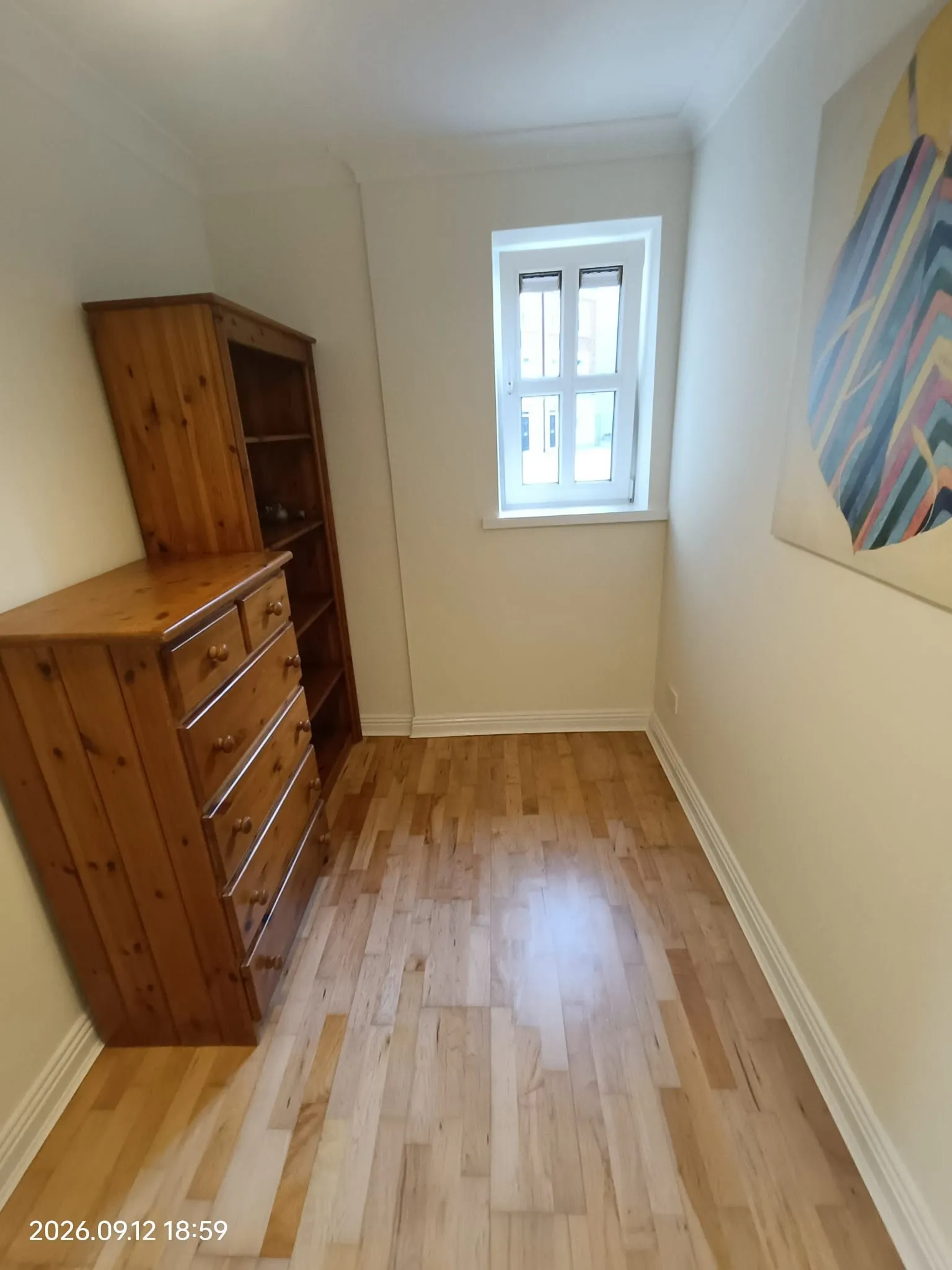 Narrow bedroom with a wooden chest of drawers and shelving unit, hardwood floor, small window, and wall art.
