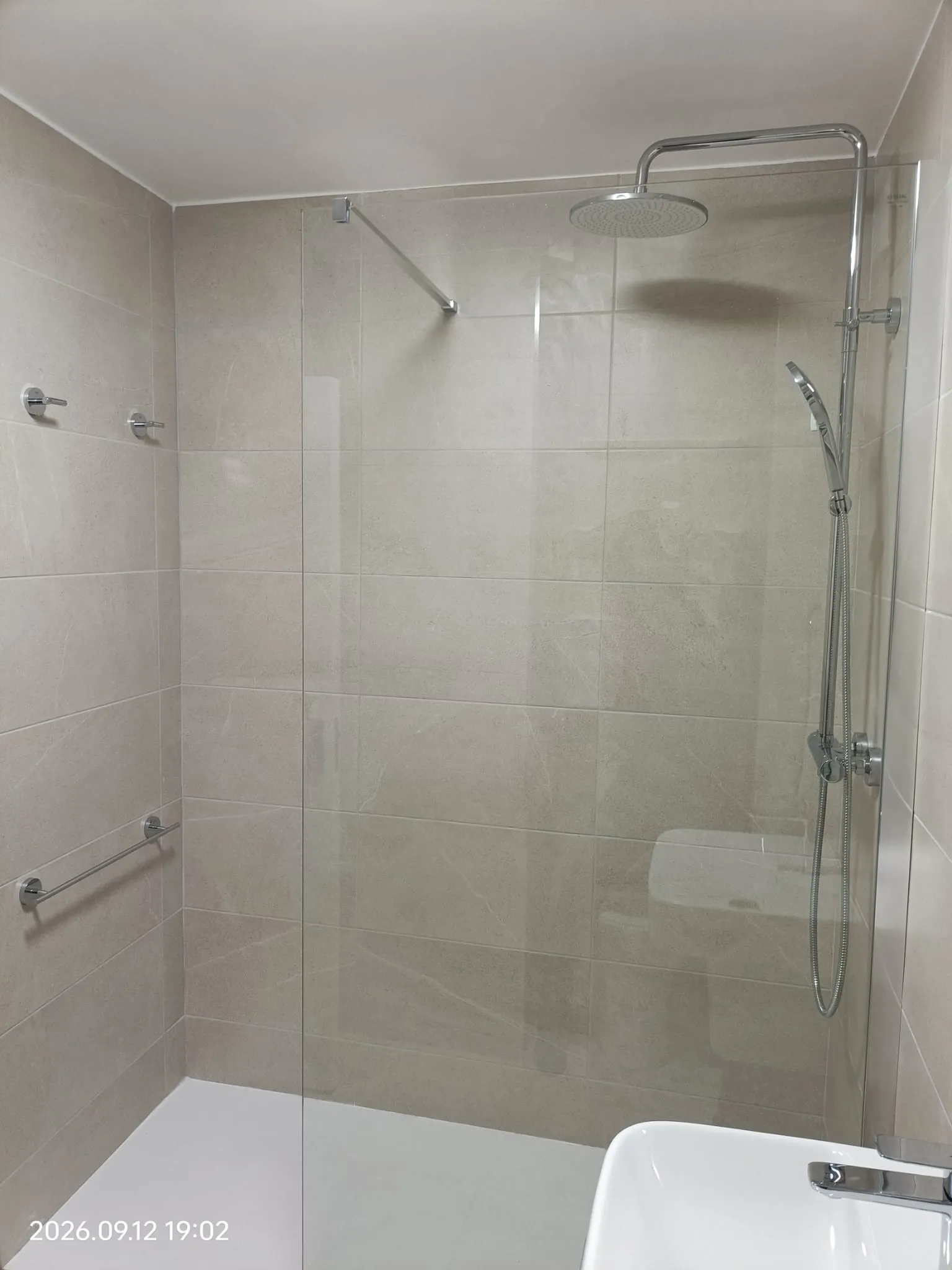 Bathroom shower area with tiled walls, glass shower screen, overhead rain showerhead and handheld shower, and a partial sink visible at the lower right