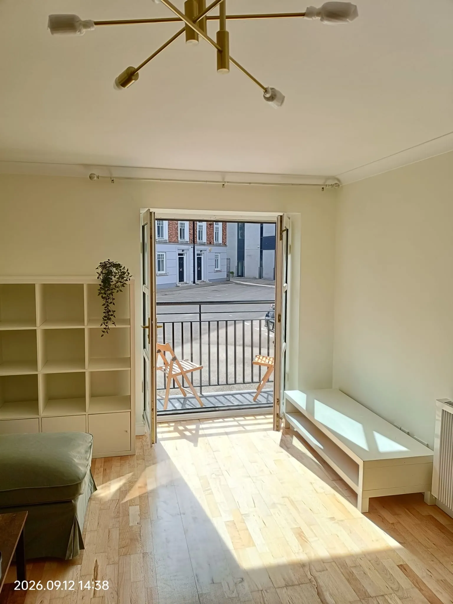 Living room with open balcony door, wooden floor, shelving unit on left, bench on right and two folding chairs visible on the balcony.