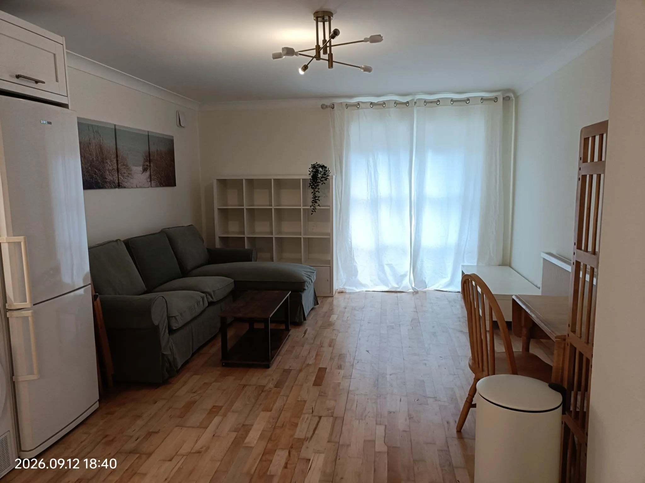 Living room with a gray sofa and chaise, a white cubed shelving unit, coffee table, dining chairs and table, a refrigerator at left, and sheer curtains covering a window.