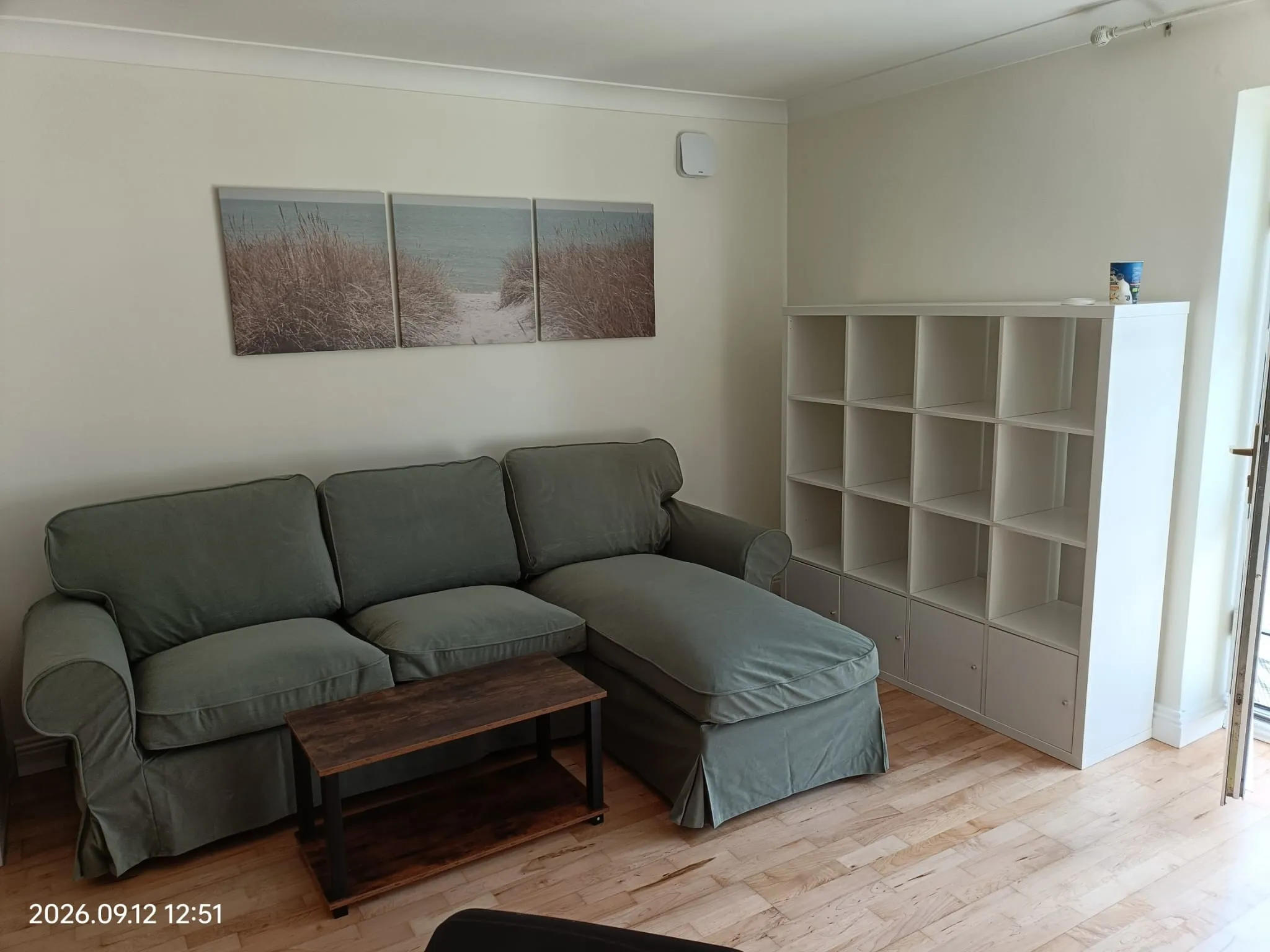 Living room with a green upholstered sofa and ottoman, wooden coffee table, white cubed shelving unit, and three-panel beach wall art on a neutral wall.