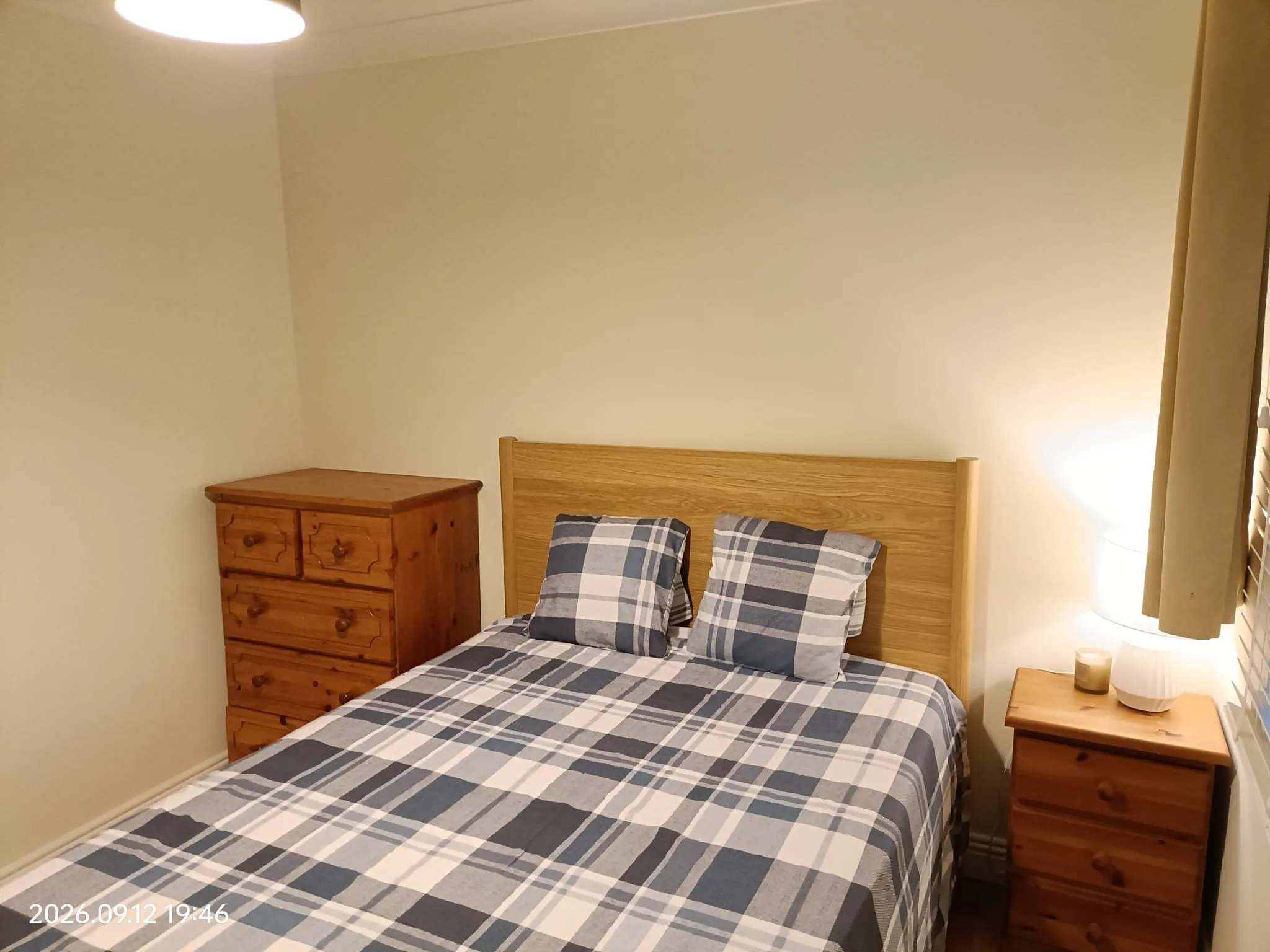 Bedroom with a double bed with plaid bedding, wooden headboard, two wooden bedside drawers and a taller wooden chest of drawers; a lamp and curtains on the right.