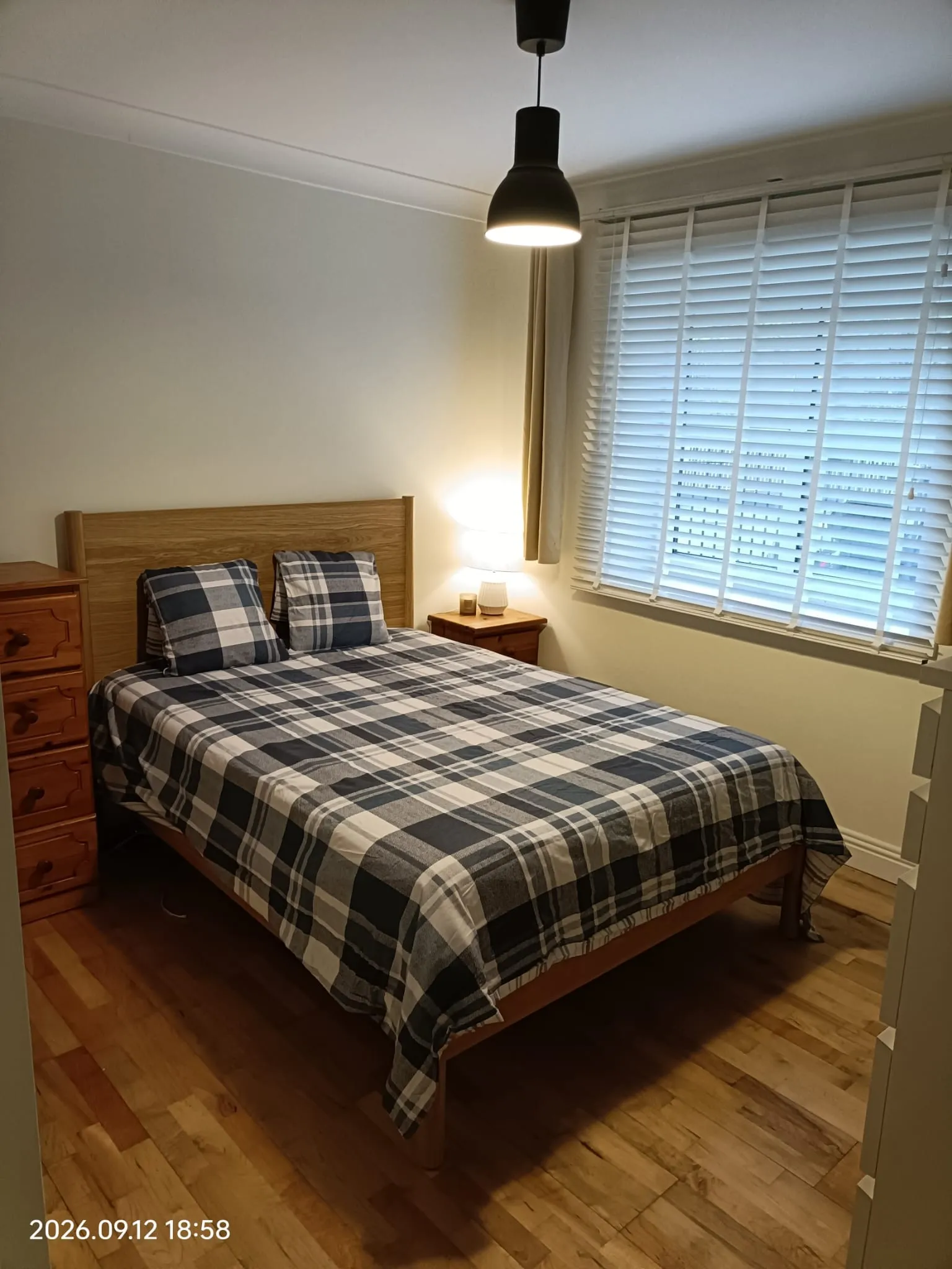 Bedroom with a double bed with plaid bedding, two pillows, wooden headboard, bedside table with a lamp, pendant ceiling light, and window with blinds.