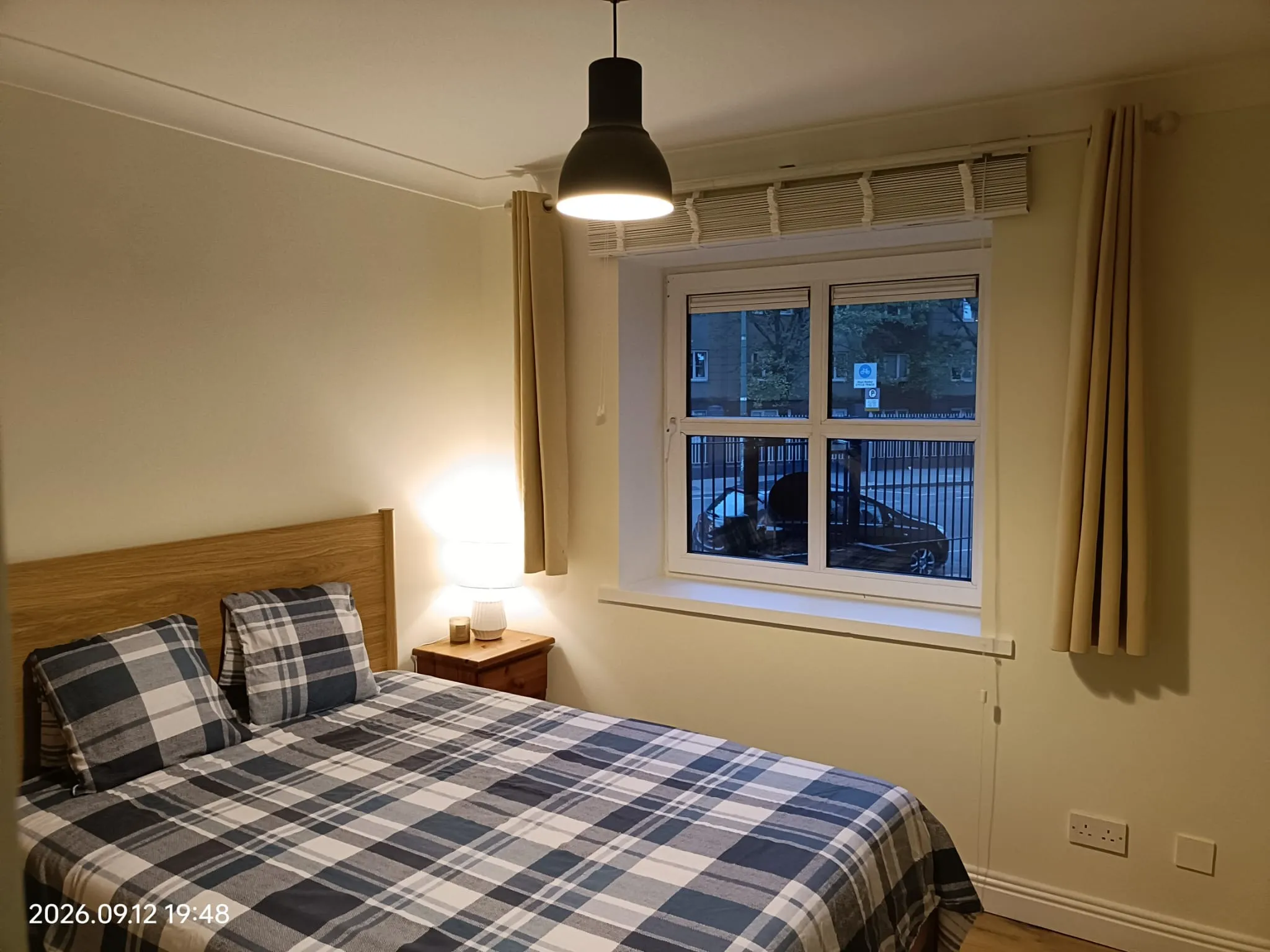 Bedroom with a double bed (plaid bedding), wooden headboard, bedside table with lamp, pendant ceiling light, and a window with blinds and curtains showing an outside street view.