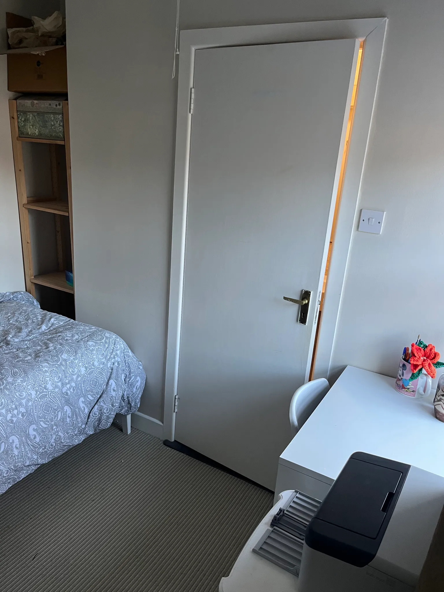 Bedroom corner with a closed white door, edge of a bed with patterned bedding, a small white desk and chair with a printer, and open wooden shelving.