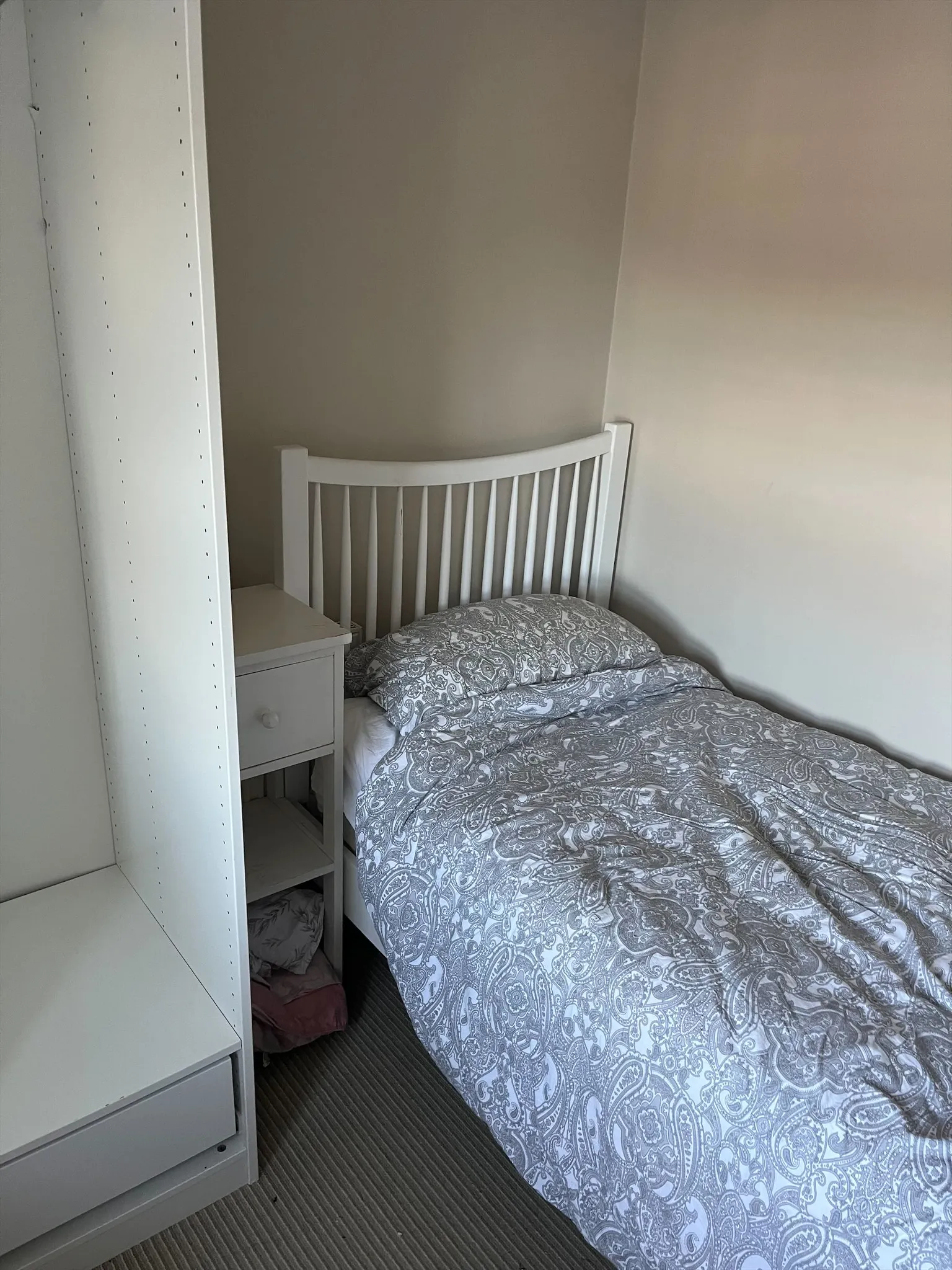 Bedroom corner with a single bed with a white slatted headboard, patterned duvet and pillow, a small white bedside table, and a tall white shelving unit